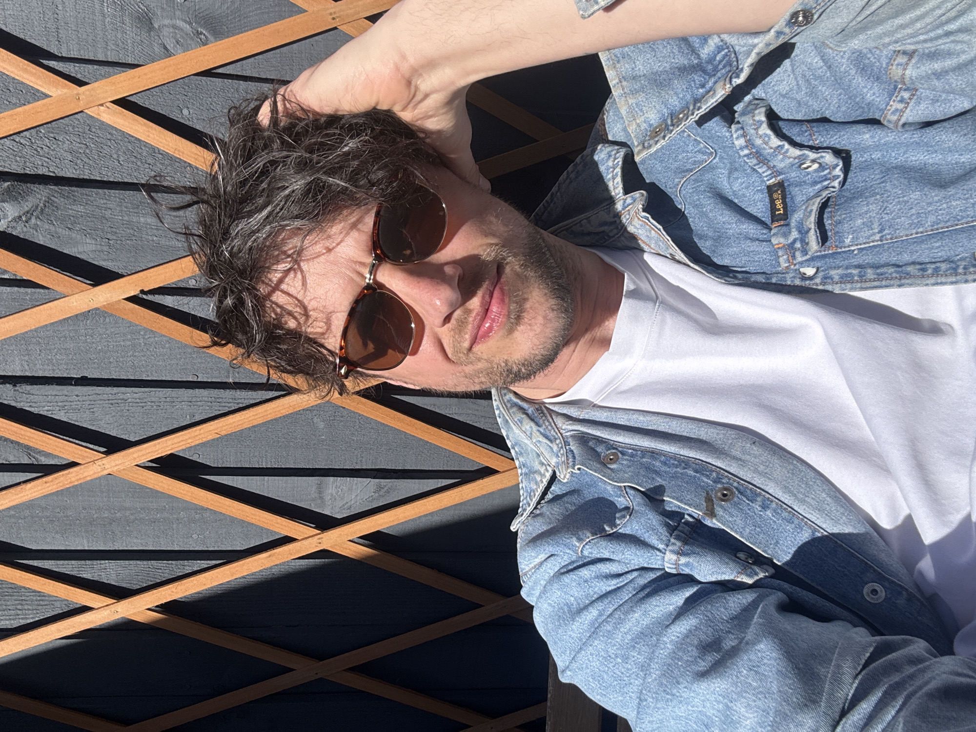 A man is pictured outdoors, possibly in a garden or patio setting. He wears sunglasses, a white t-shirt, and a denim jacket. He is positioned in front of a wooden structure, possibly a fence or decorative trellis, with an alternating criss-cross pattern. The structure appears to be painted in a dark color, possibly gray or blue-gray. The lighting suggests a sunny day, casting distinct shadows on the background.
The image provides insufficient information to specify a precise location within South Kesteven, United Kingdom.