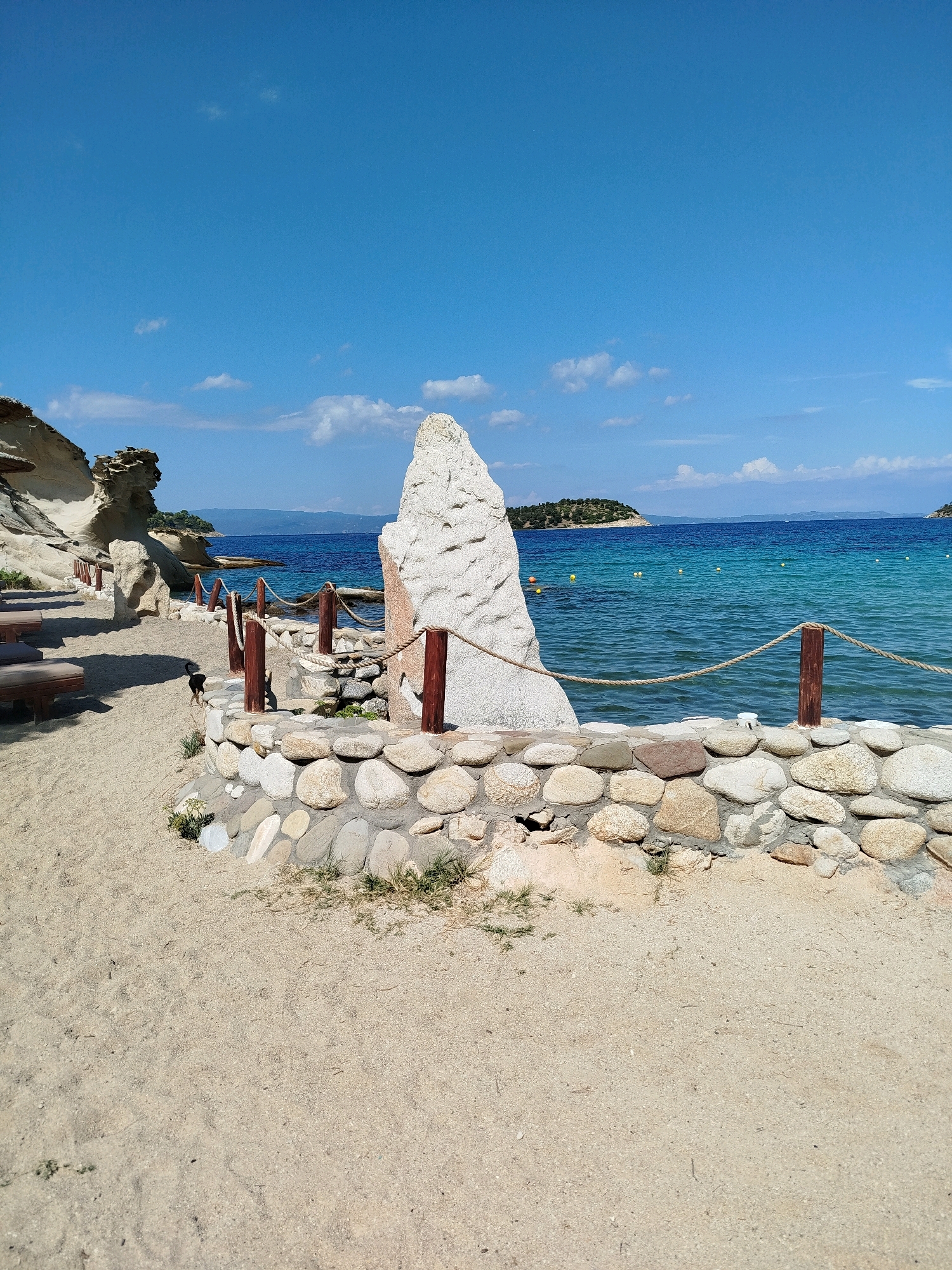 The image depicts a serene beach scene in Sithonia Municipal Unit, Greece, with a clear blue sky overhead. The foreground is dominated by a sandy beach, with a low stone wall bordering a section of the sand. A rope railing, supported by wooden posts, runs along the top of the stone wall. To the left of the stone wall, beach loungers are visible on the sand, casting shadows. A small black dog is walking on the sand in the middle ground. Behind the stone wall, the turquoise water of the sea stretches out to the horizon. In the distance, small green islands dot the sea, and buoys are scattered on the water. The overall impression is one of a tranquil, sun-drenched coastal landscape.