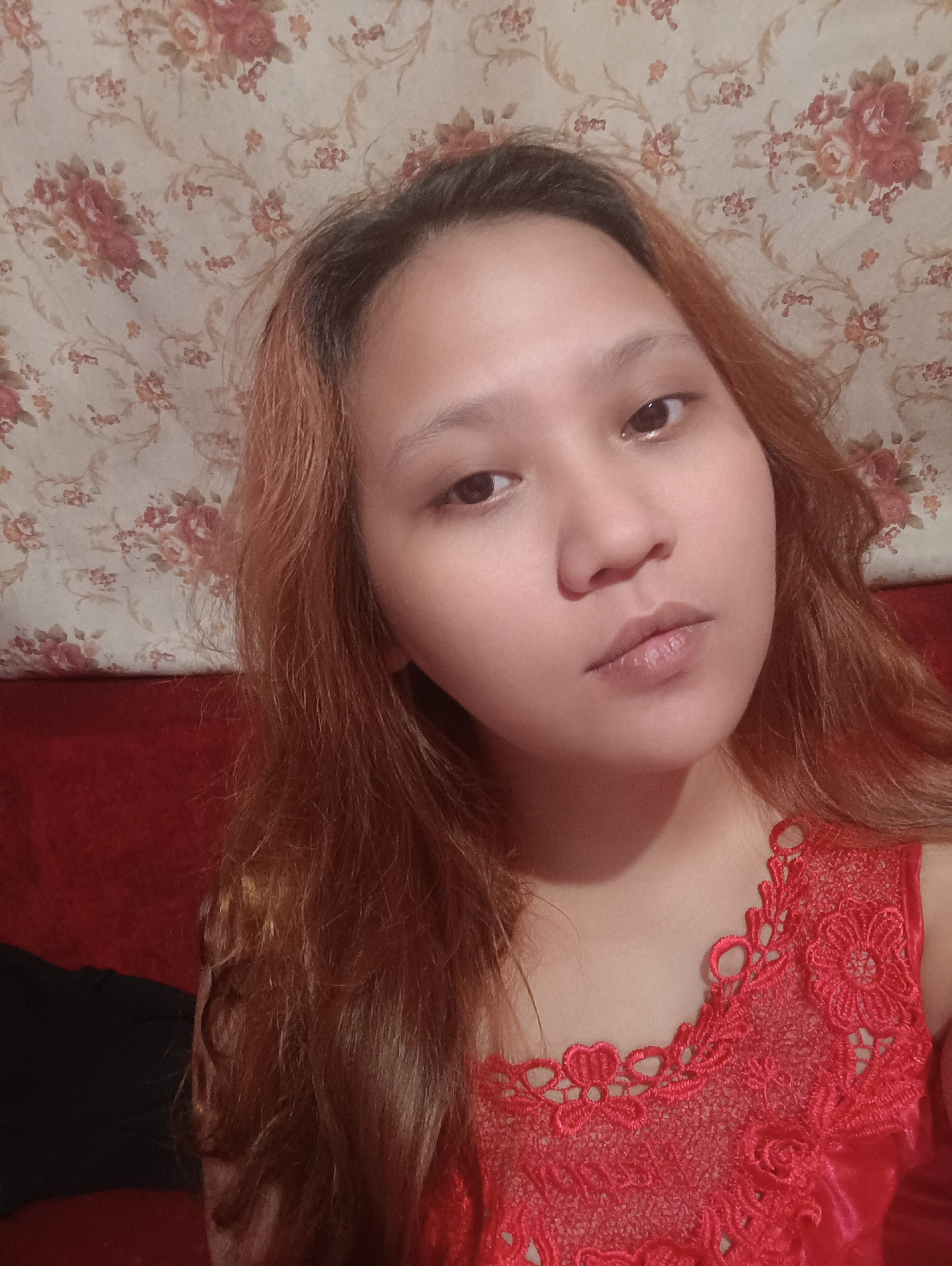 A young woman with long, reddish-brown hair is looking directly at the camera. She is wearing a red lace top. She is sitting in front of a floral patterned wallpaper and a red sofa. The location is Depok, Indonesia.