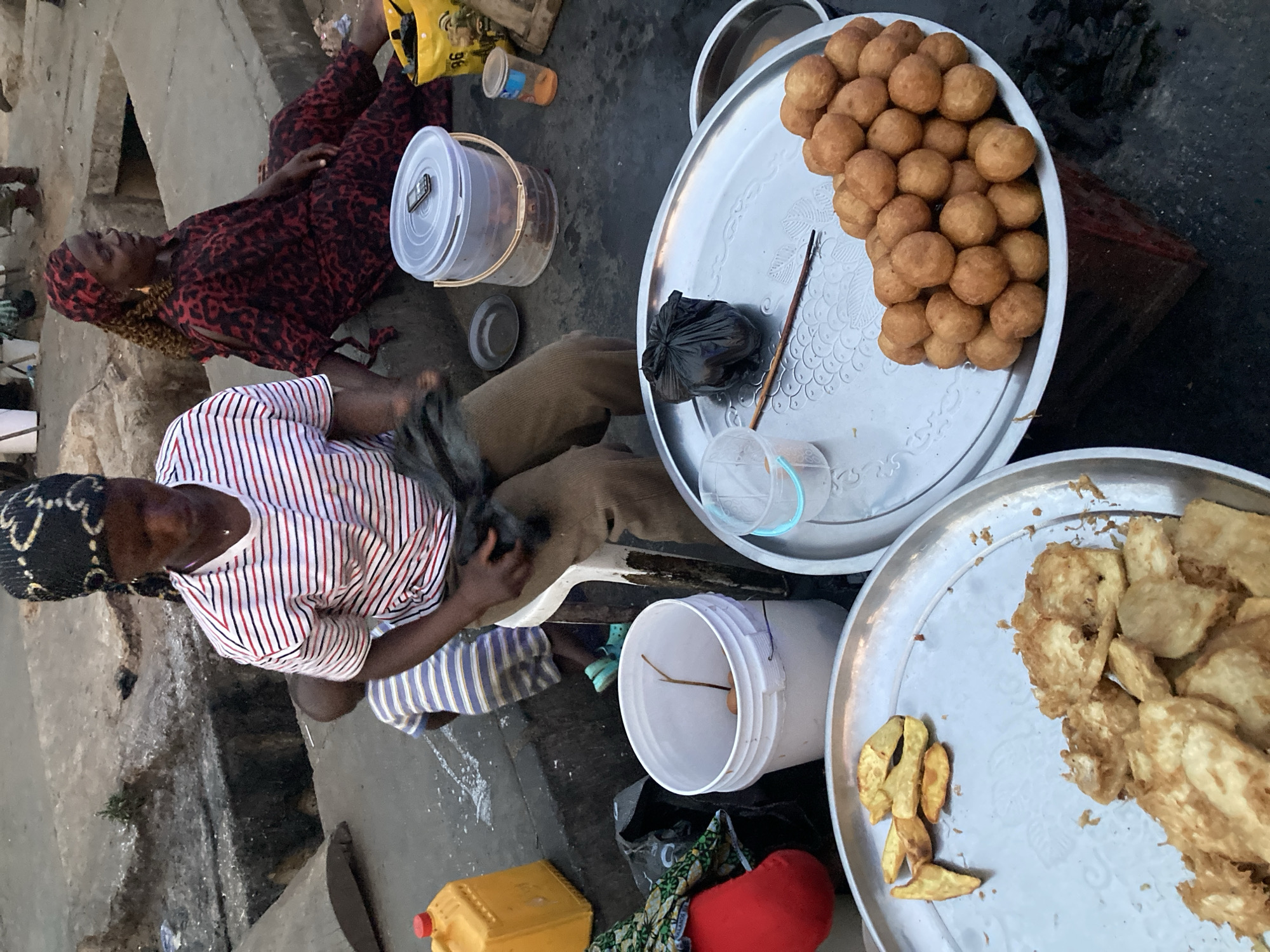 The image shows a street food vendor in Jos, Nigeria. Two women are visible, one sitting on the ground in a red and black patterned dress, and the other crouched nearby, wearing a striped shirt and black head covering. In front of them are three large metal trays displaying prepared food. One tray contains small, round, fried balls, while another has fried plantain chips. A white bucket, plastic containers, and a yellow jerrycan are also present, along with other scattered items. The background shows a weathered concrete surface and elements of a typical urban street.