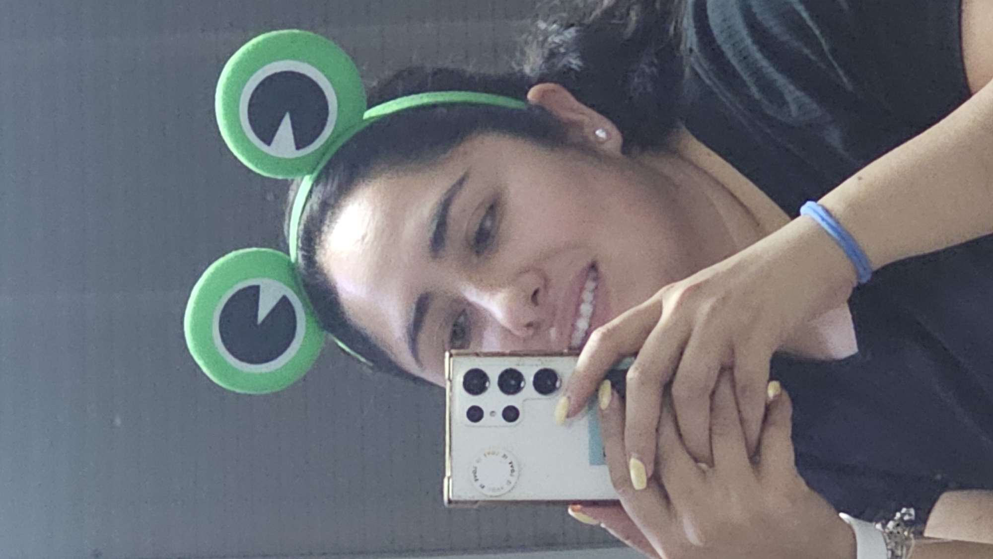 A young woman is taking a selfie in a mirror, possibly in San Bernardino Tlaxcalancingo, Mexico. She is wearing a green frog-themed headband and smiling. She is holding a white smartphone with multiple rear-facing cameras. She has pale yellow nail polish. The woman is wearing a black shirt. On her wrist she has a blue bracelet. She is also wearing a silver wristwatch with a white band.