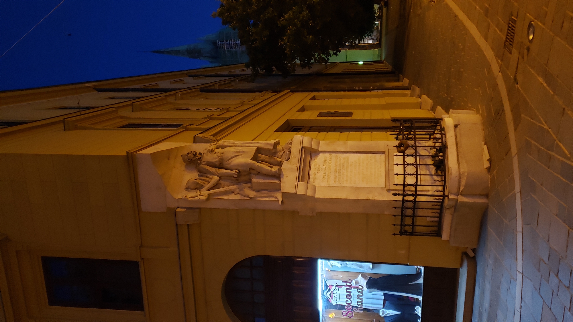 The image depicts a corner of a building in the city of Prešov, Slovakia, at dusk. The building is light yellow and has multiple windows. On the corner, there is a stone sculpture of two figures, one standing and one seated, with a plaque below. A black iron fence surrounds the base of the sculpture. To the left, a shop window displays clothing and mannequins, with a sign that reads "LUXUS Second hand". A cobblestone sidewalk is visible in the foreground, and a street extends into the background. In the distance, the spire of a green-roofed church is visible against the dark blue sky. The lighting suggests it is nighttime, with artificial lights illuminating the storefront and parts of the building and sidewalk.