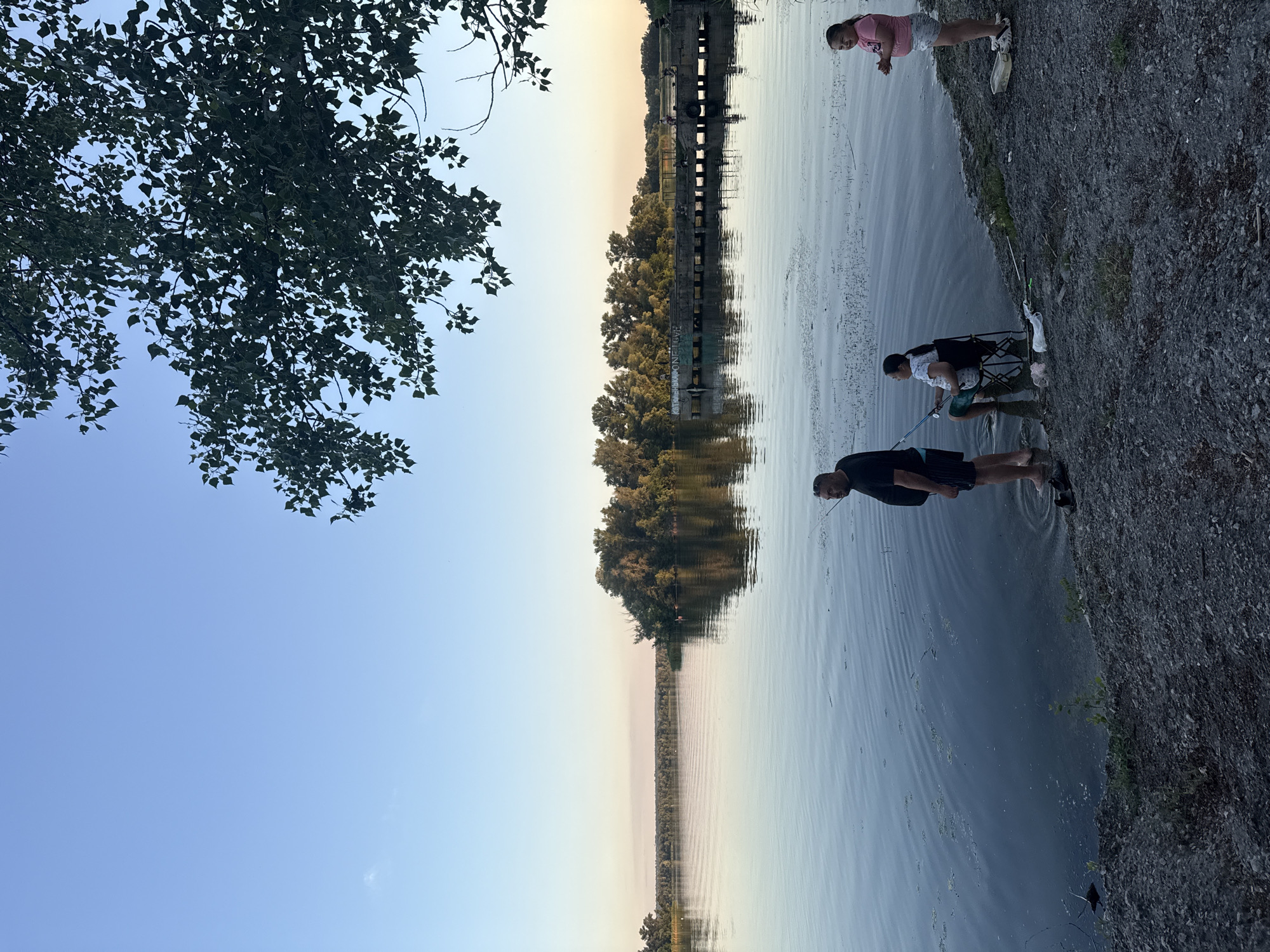 In Obukhivka, Ukraine, a group of people are engaged in fishing activities by a wide, calm river. The scene is set during the late afternoon, with a clear blue sky transitioning to softer hues near the horizon, where a line of trees and a distant structure are silhouetted. On the right bank, a man stands near the water's edge, facing the river. Beside him, a woman is seated in a folding chair, actively fishing with a rod. A young girl stands a bit further to the right, observing the activity. The riverbank is covered in small stones and gravel, with some sparse vegetation. A pier or bridge extends into the water in the middle distance, with several figures visible on it. Lush green trees form a dense backdrop on the far bank, reflecting on the water's surface. The overall atmosphere is serene and recreational.