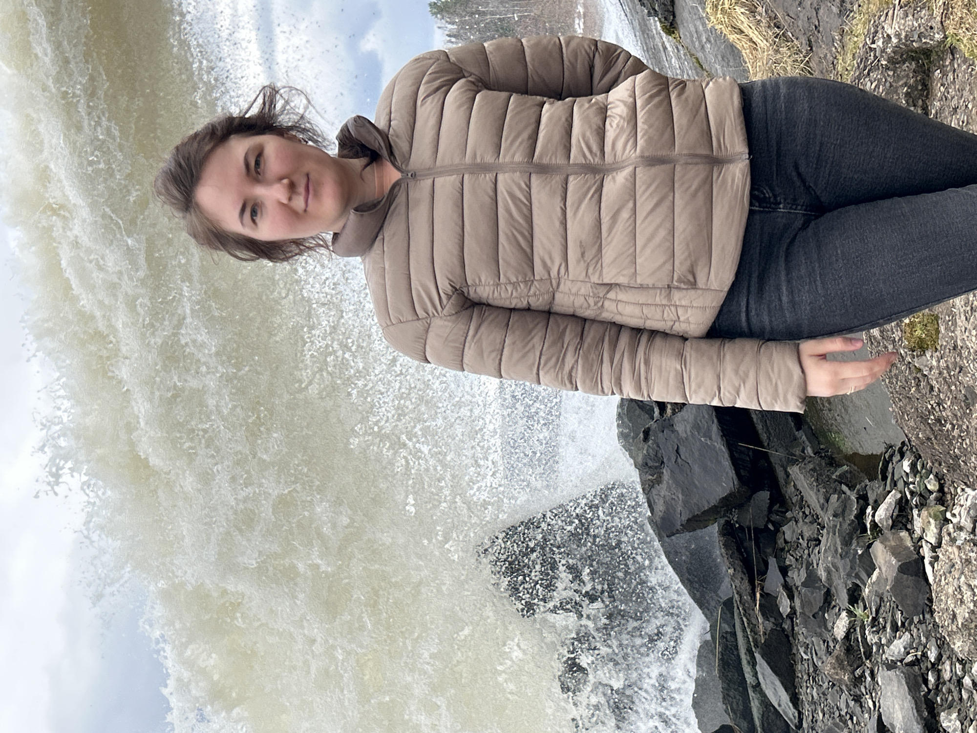 A medium shot depicts a woman standing in front of a waterfall. The woman is wearing a tan puffer jacket and dark jeans. She is looking at the camera. The waterfall fills much of the left side of the frame. Black rocks are visible at the bottom right of the frame. The image was taken in the city of Chernushka, Russia.