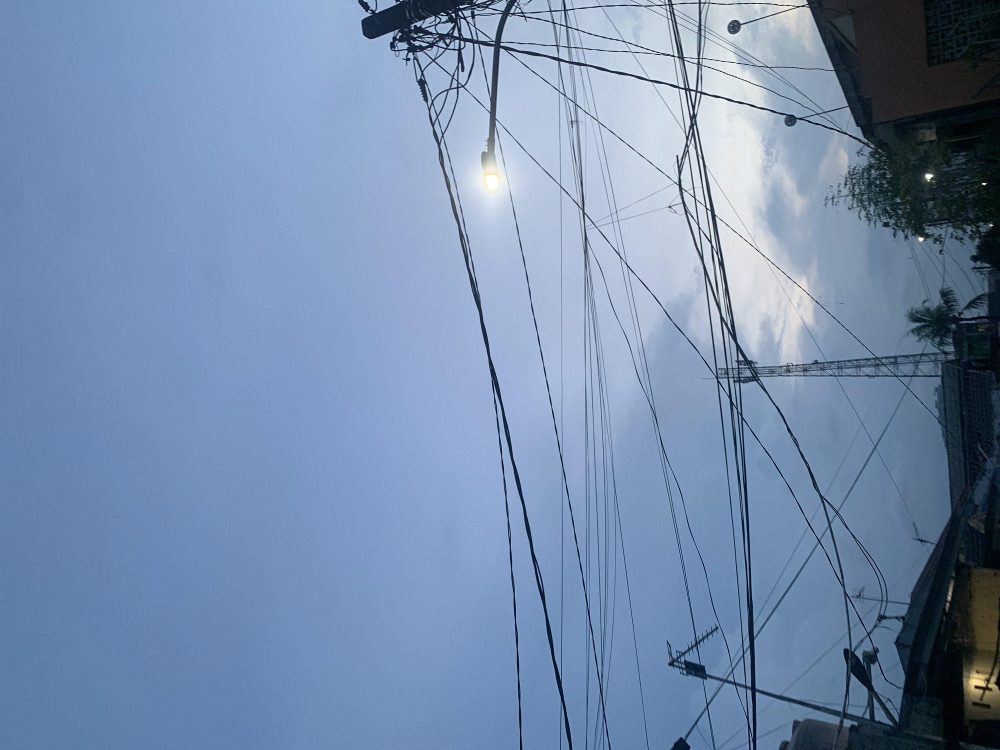The photograph, taken in the city of Bekasi, Indonesia, shows a low-angle view of a daytime sky crisscrossed with power lines and communication cables. The sky transitions from a clear blue on the left to a cloudy, overcast area on the right. A street lamp is visible, illuminated despite the daylight. Building structures are partially visible on the right side of the frame. A tall communication tower rises in the background, partially obscured by clouds and foliage.