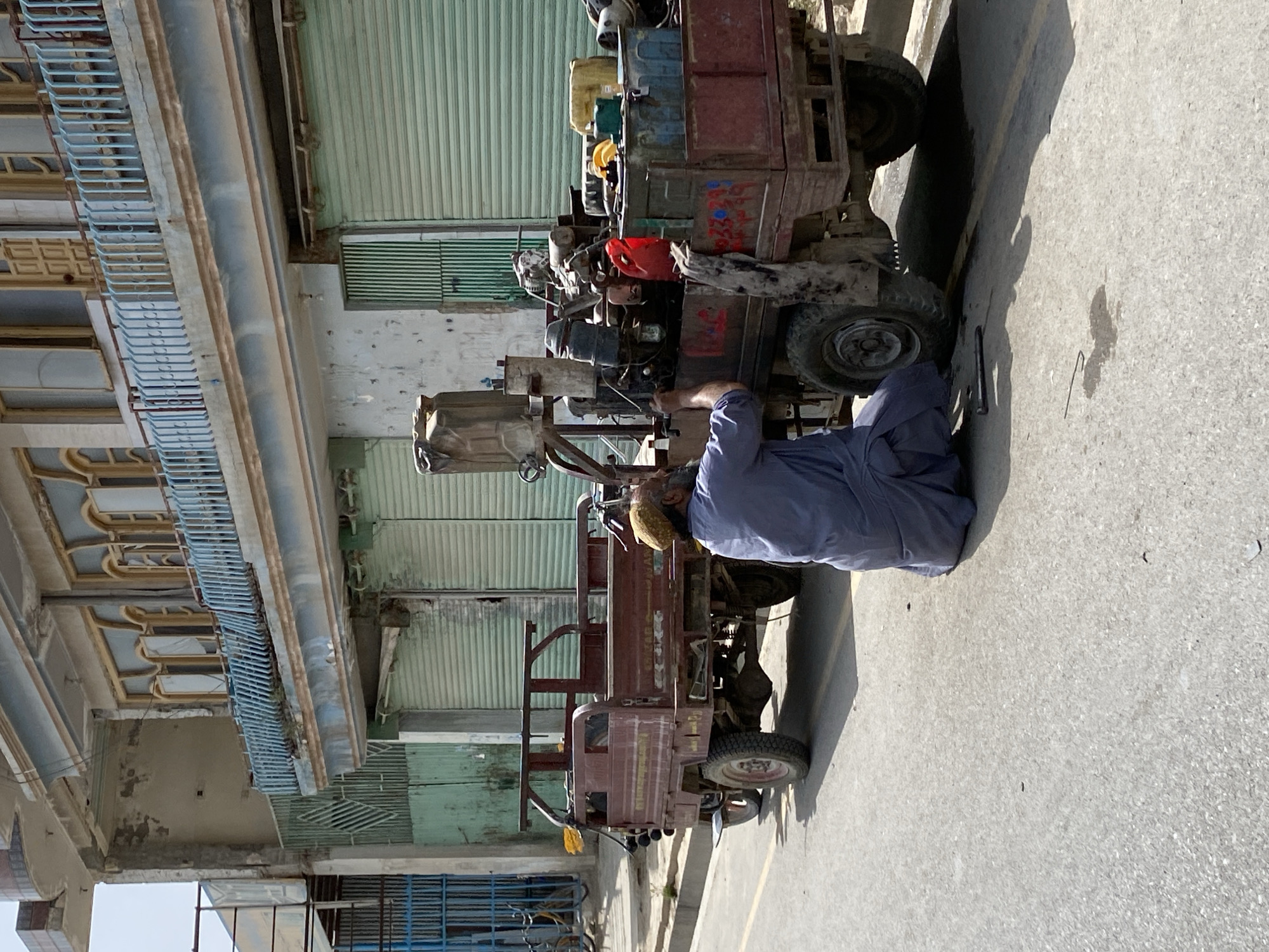 A man wearing a blue shalwar kameez and a patterned cap is working on a three-wheeled vehicle that appears to be a generator or water pump mounted on a flatbed truck. The vehicle is parked on the side of a paved road in front of a building with several closed roll-up doors and an upper-level balcony with a blue railing. The building's facade is weathered, with peeling paint and some visible damage. The vehicle is red and appears to be loaded with various mechanical parts, containers, and a red jerrycan. A tool, possibly a wrench, lies on the ground near the man's feet. The scene is outdoors, under daylight, with a clear sky. The overall impression is of a street-side repair in an urban environment in Kandahar, Afghanistan.