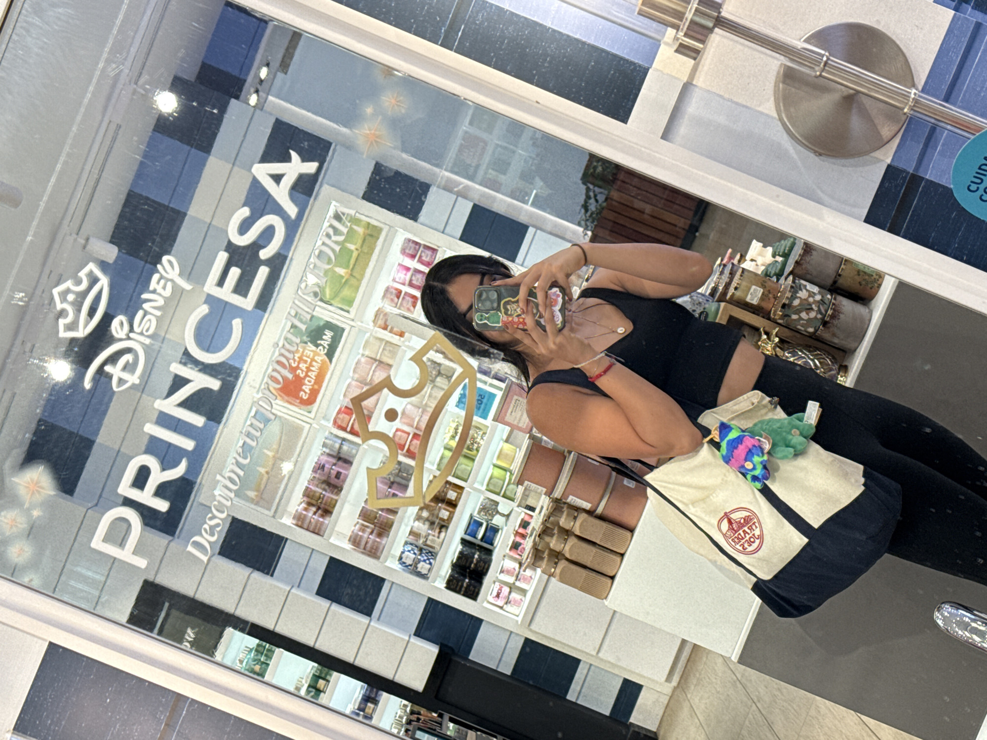 A photograph captures a person reflected in a storefront window in Puebla, Mexico. The store, "Disney Princesa," has its name prominently displayed in white lettering on the glass. The person in the reflection is holding a phone up to take a photograph. They are wearing a black tank top and black leggings, and they have a tote bag over their shoulder. Inside the store, there are shelves stocked with various items, possibly candles or other merchandise. The storefront has a white and blue checkered pattern below the window.