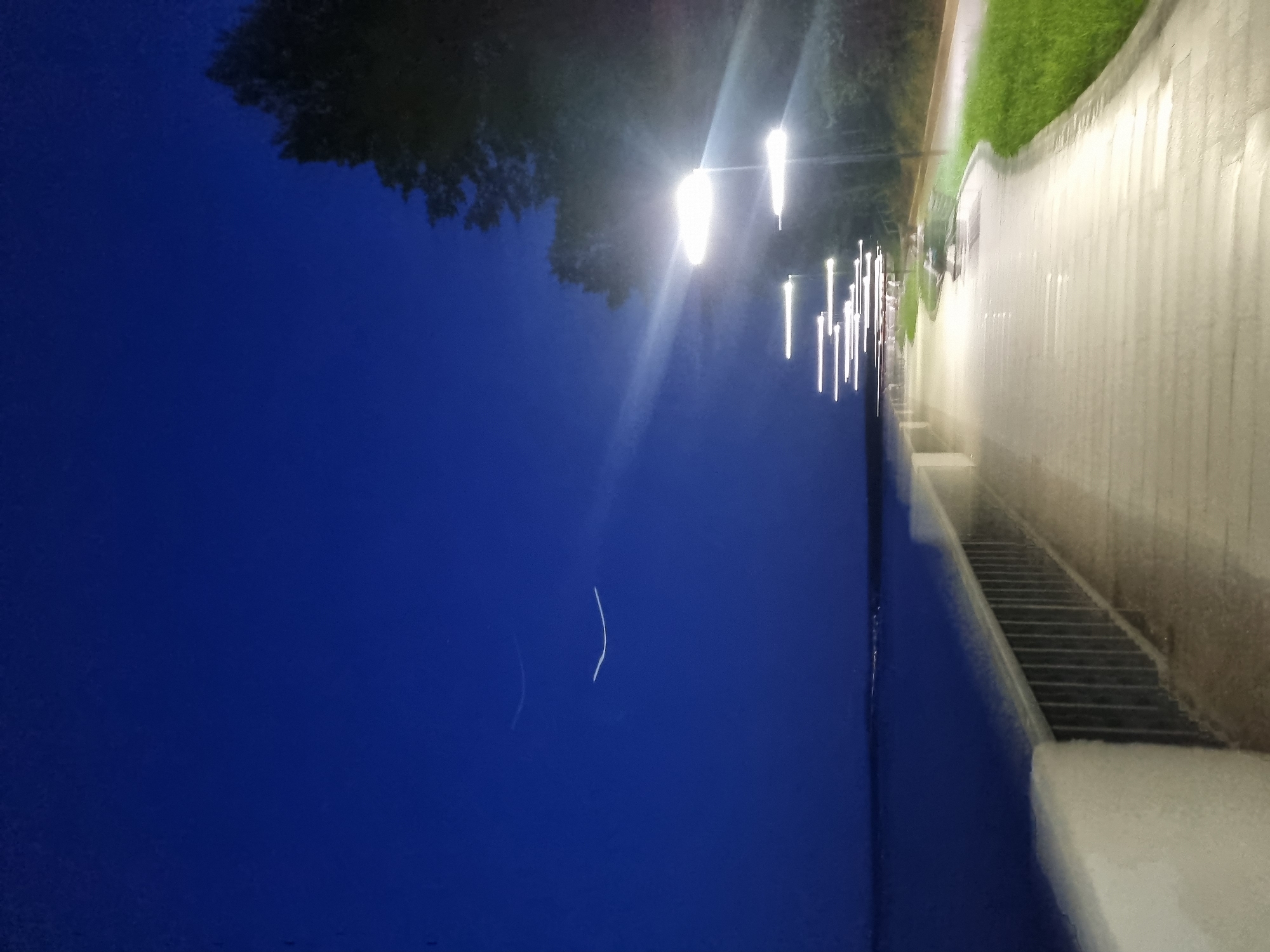 At night, a view of the embankment in Chaikovsky, Russia shows numerous bright streetlights illuminating the path along the shoreline. A series of steps lead down to the water. The sky is a deep blue, and trees are visible in the background.