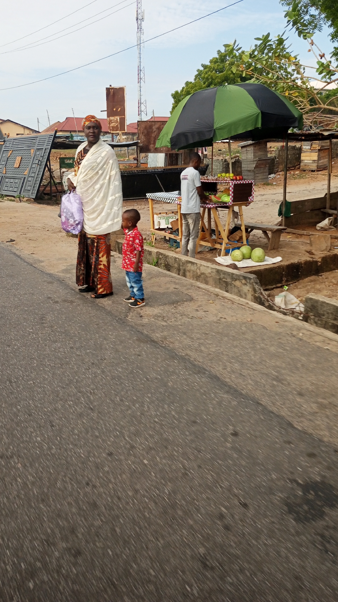 The image depicts a street scene in Lokoja, Nigeria. An adult female wearing traditional attire and carrying a purple bag stands on a sidewalk next to a young child in a red shirt and jeans. Adjacent to them is a fruit stand covered by a green and black umbrella where a man is arranging fruit. Three watermelons are placed on a white cloth by the fruit stand. Metal gates are visible in the background, and a cell tower is prominent in the distance. The immediate surroundings include the paved road and roadside structures.