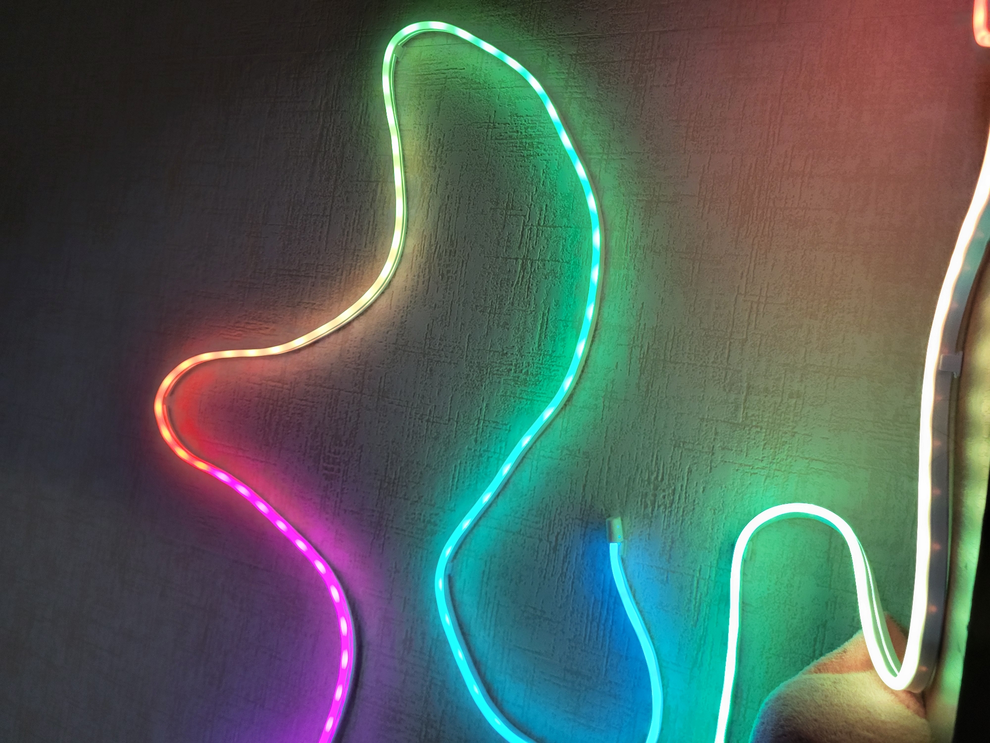 A string of colorful LED lights is draped across a textured, dark surface, possibly a tabletop or wall. The lights transition through a gradient of colors, starting with purple and pink on the left, moving through red and orange, and then fading into green and blue on the right. The LED string is illuminated, creating a soft glow that reflects on the surface. The surface itself has a subtle pattern of lines or grooves. In the lower left corner, a small portion of a light pink, fuzzy object is visible, which may be a piece of fabric or material. The scene appears to be indoors, with no discernible elements suggesting a specific outdoor location in Horishni Plavni, Ukraine.