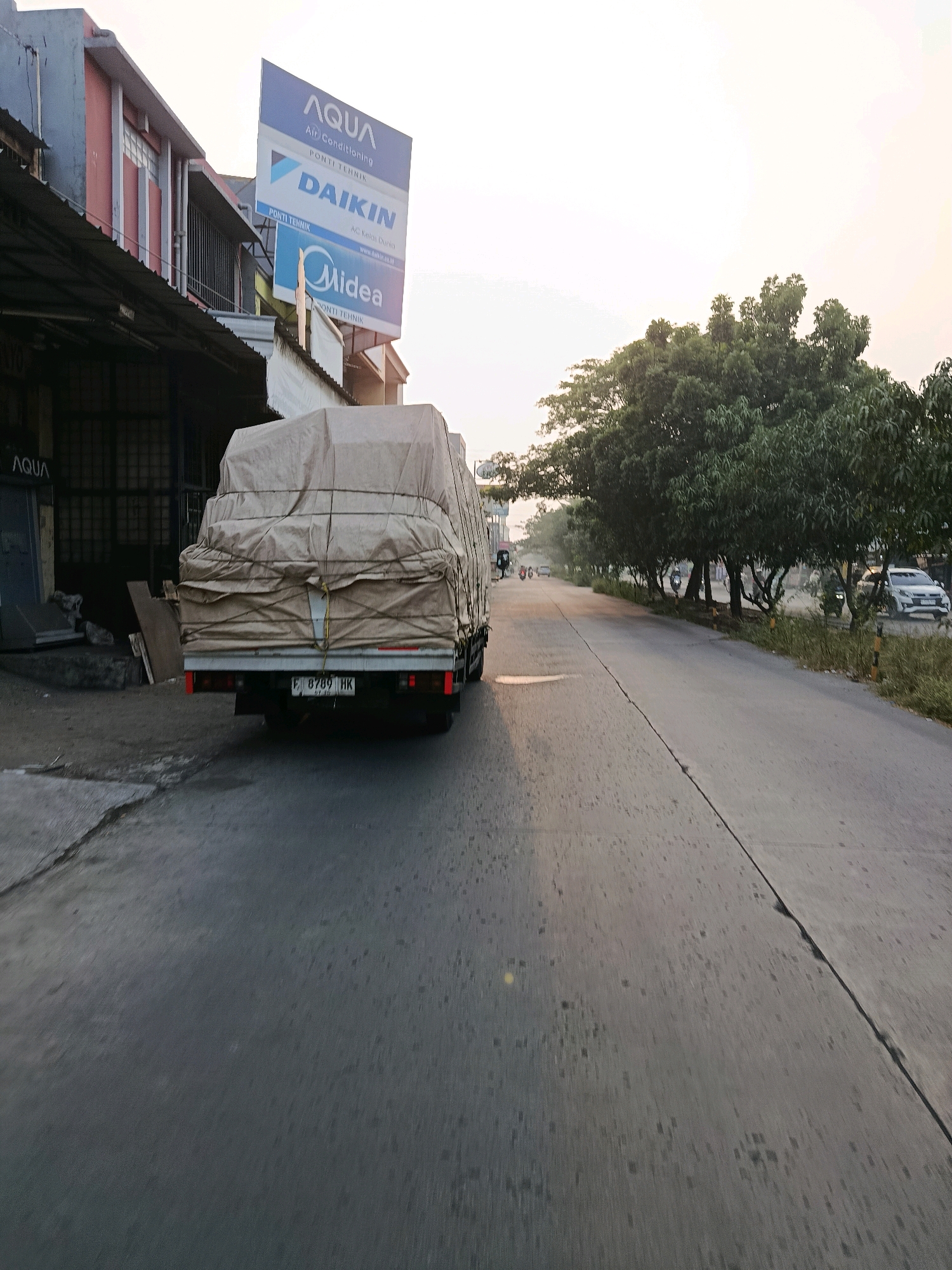An exterior, eye-level, wide shot captures a street scene in Bekasi, Indonesia, at dusk. The sky is a pale orange, transitioning into white. On the left, a building with a gray and pink facade is partially visible, with a dark awning over a storefront. A large billboard advertising AQUA Air Conditioning, DAIKIN, and Midea is mounted on the building. In the foreground, a light brown tarp-covered truck is parked. The truck has a license plate reading "B 8789 HK". The road is asphalt and cracked, with grass and trees lining the right side. Several vehicles and motorbikes are present on the road, and two figures, possibly pedestrians, are walking in the distance.
