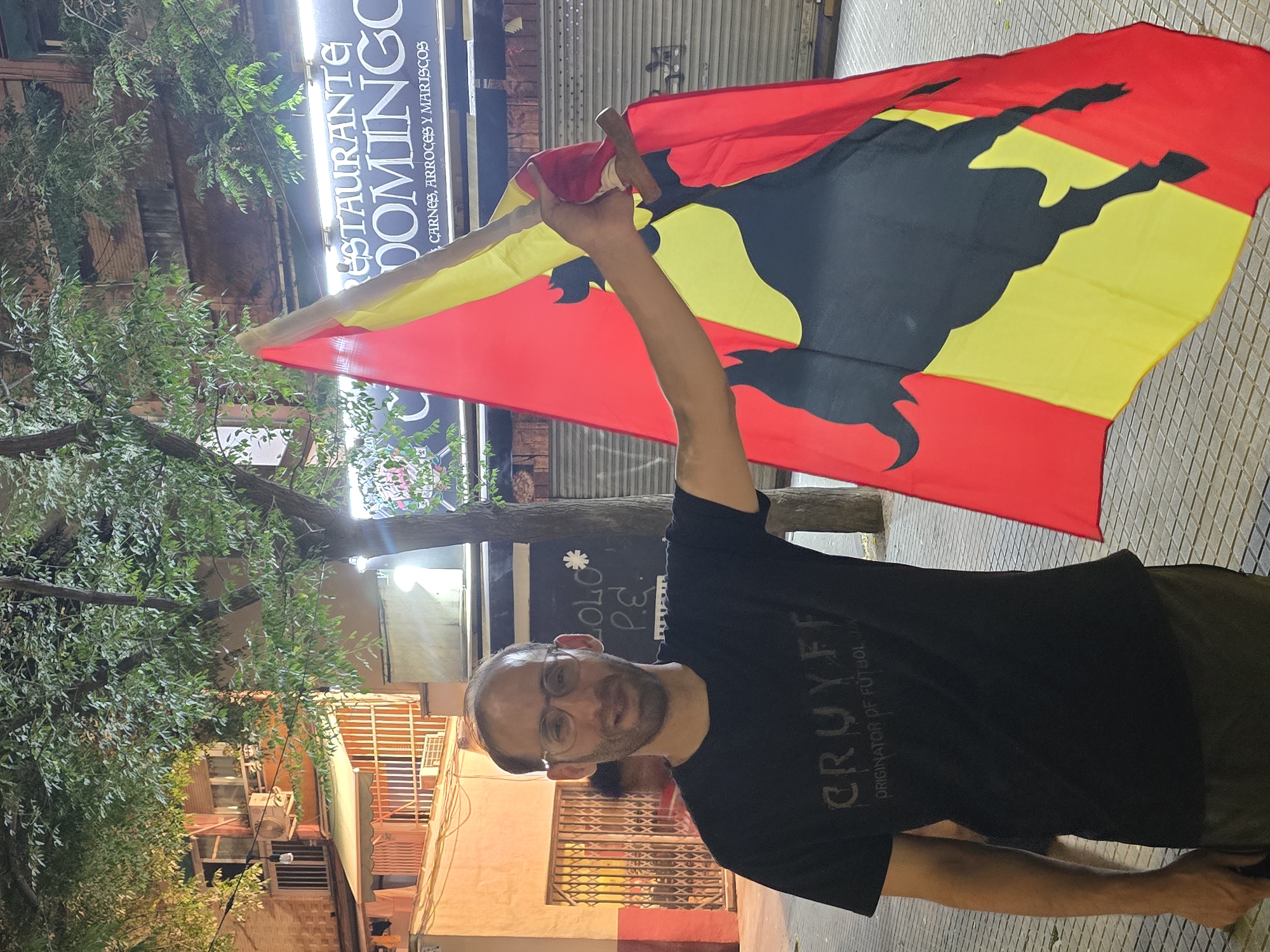 A man with glasses and a beard, wearing a black t-shirt with the text "CRUYFF ORIGINATOR OF FÚTBOL" printed on it, stands holding a flag with a black bull silhouette on a background of red and yellow stripes. He is positioned on a tiled sidewalk in front of a building. To his right, a sign reads "RESTAURANTE DOMINGO" followed by "CARNES, ARROCES Y MARISCOS." The background includes trees, building facades with windows and balconies, and streetlights. The scene appears to be outdoors at night, possibly in a commercial or residential area of Cornellà de Llobregat, Spain.