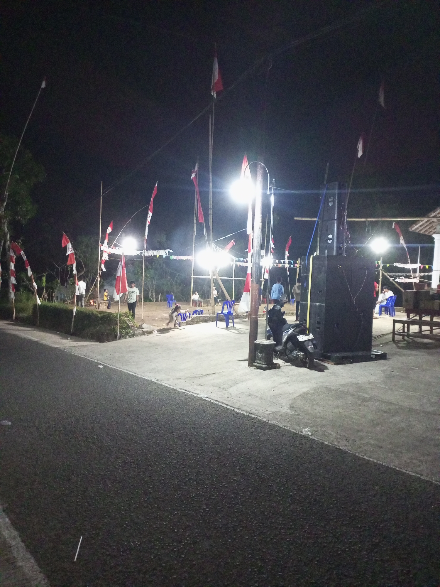 This image depicts an outdoor scene at night, likely in Jatipurno, Indonesia, given the presence of Indonesian flags. The foreground shows a paved area adjacent to a road. A group of people is gathered in the middle ground, some standing, some sitting. Red and white Indonesian flags are prominently displayed on bamboo poles throughout the area, indicating a possible celebration or event. In the background, trees and foliage are visible against a dark sky illuminated by artificial lights. A large speaker system is set up on the right side of the image, suggesting a musical or public address event. A motorcycle is parked in front of the speaker system. Several blue plastic chairs are arranged in small groups, with some people seated in them. The overall atmosphere appears festive or communal.