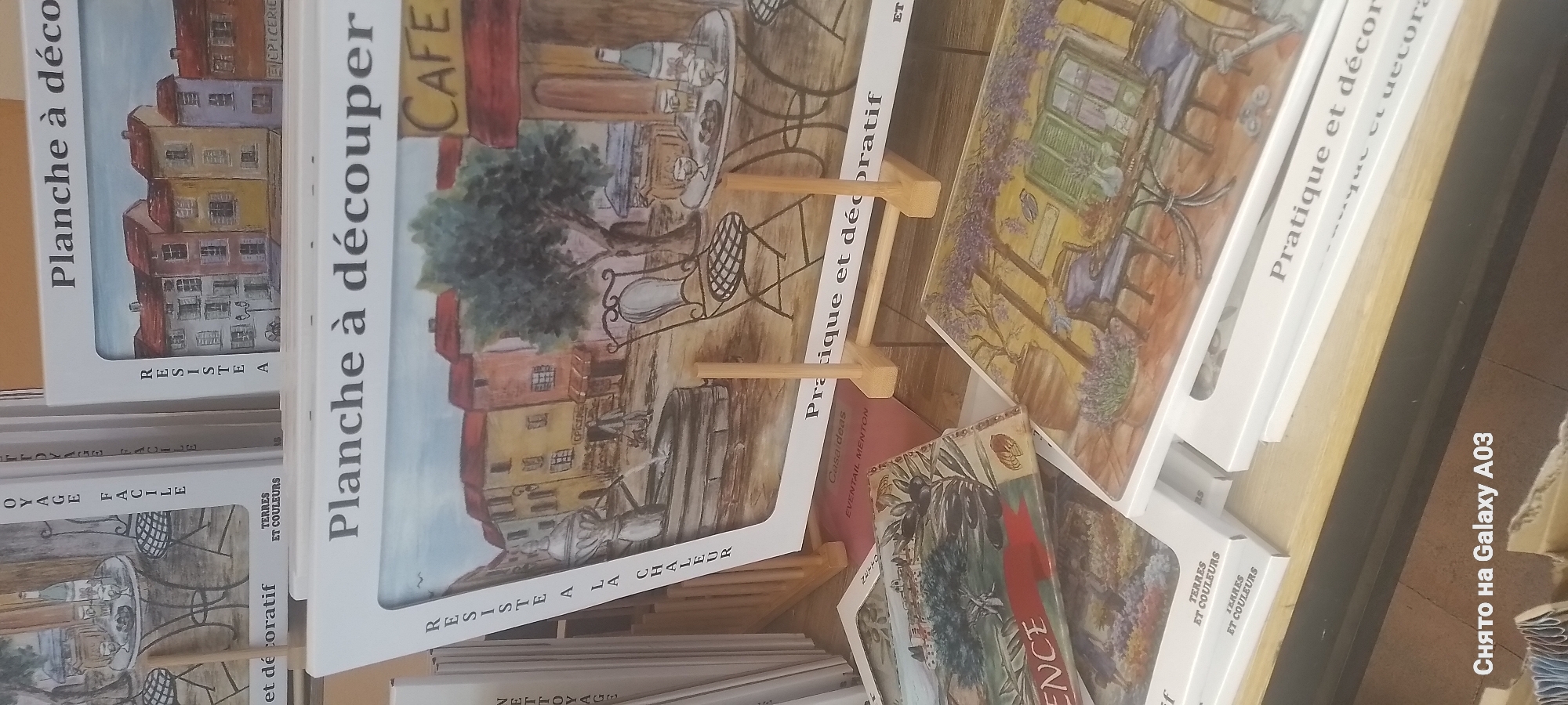 The image shows a display of decorative items, likely ceramic tiles or placemats, in what appears to be a shop or market stall. The items feature watercolor-style illustrations of French scenes. One prominent item is labeled "Planche à découper" and depicts a street scene with buildings and a cafe. Other items display images of lavender fields and olives, evoking the Provence region. The text "TERRES ET COULEURS" is visible on the packaging of some items. The bottom of the image includes a watermark in Russian, "Снято на Galaxy A03" which translates to "Shot on Galaxy A03", indicating the device used to capture the photograph. The overall aesthetic suggests a souvenir or decorative art vendor, possibly catering to tourists interested in the French Riviera, with a specific mention of "Menton" on one of the items ("EVENTAIL MENTON"). The lighting is consistent with indoor retail display lighting. There are no people or animals visible in the frame.