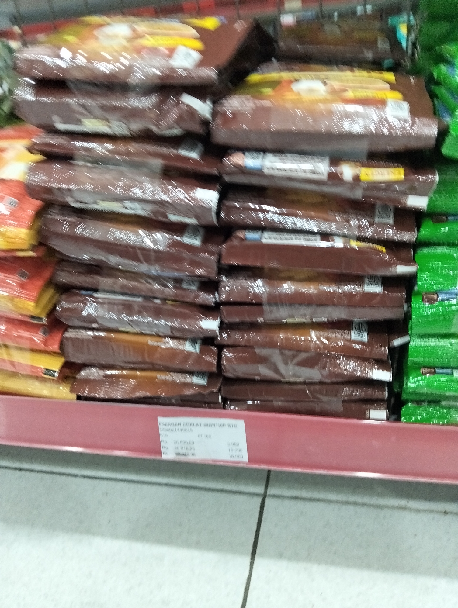 The image shows a retail shelf in a store. Stacked on the shelf are numerous individually wrapped chocolate bars. The wrappers are primarily a dark brown color, with some displaying yellow and white accents. To the right of the chocolate bars, there are green packaged items, also stacked. A red shelf edge is visible in the foreground. A price tag is attached to the red shelf edge, displaying text that includes "ENERGEN COKLAT 29GR 10P RTG" and monetary values like "Rp. 20.500.00" and "Rp. 20.219.96". The floor beneath the shelf appears to be tiled with light-colored squares. The image quality is somewhat blurry, suggesting it was taken with a mobile device or in low light conditions. The context provided indicates this scene is located in the city of Kedungrejo, Indonesia.