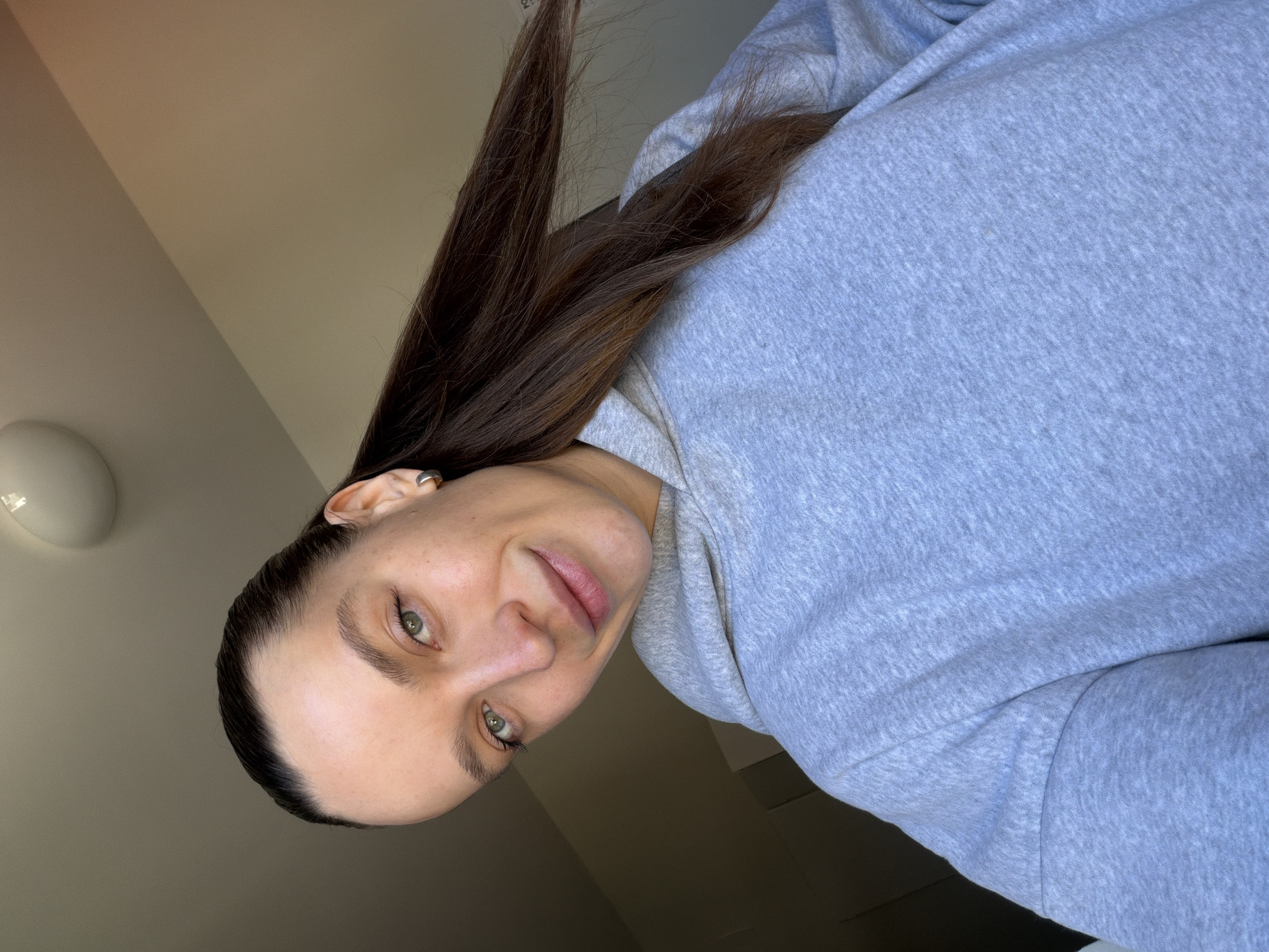 A woman with fair skin, light green eyes, and dark brown hair is wearing a gray hooded sweatshirt. The image is taken from a low angle, slightly rotated, showing her face, hair falling over her shoulder, and part of the sweatshirt. The background consists of a plain, light-colored wall with a round light fixture. There is no location information pertaining to Budva, Montenegro, discernable from the image.