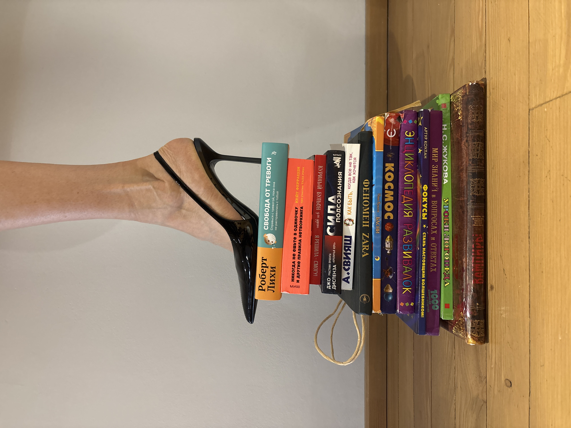 A close-up, eye-level shot captures a bare leg and foot, extended horizontally from the left, with the foot clad in a shiny black high-heeled shoe. The slender heel of the shoe precariously rests on the top edge of the first book in a vertical stack. The scene is set indoors against a plain, light-colored wall in the background and a warm-toned wooden floor with visible grain on the right.
The arrangement of the foot and books suggests an artistic or conceptual photograph, possibly emphasizing balance or a juxtaposition of elegance and intellect. The books, varying in size, color, and binding style, are predominantly Russian titles. Prominently visible texts include "Роберт Лихи" (Robert Leahy) and "СВОБОДА ОТ ТРЕВОГИ" (Freedom from Anxiety) on an orange book. Other titles visible are "НИКОГДА не ешьте в ОДИНОЧКУ" (Never Eat Alone), "СИПА ПОДСОЗНАНИЯ" (SIPA Subconscious), "ФЕНОМЕН ZARA" (Zara Phenomenon), "КОСМОС" (Cosmos), "ЭНЦИКЛОПЕДИЯ РАЗВИВАЛОК" (Encyclopedia of Developables), and "МИР ЗНАНИЙ В ВОПРОСАХ И ОТВЕТАХ" (World of Knowledge in Questions and Answers). A dark, ornate-bound book at the end of the stack might be titled "ВАМПИРЫ" (Vampires). A thin, braided rope or string is draped near the base of the book stack, adding a subtle detail. The lighting is soft and even, highlighting the textures of the skin, shoe, and book spines.