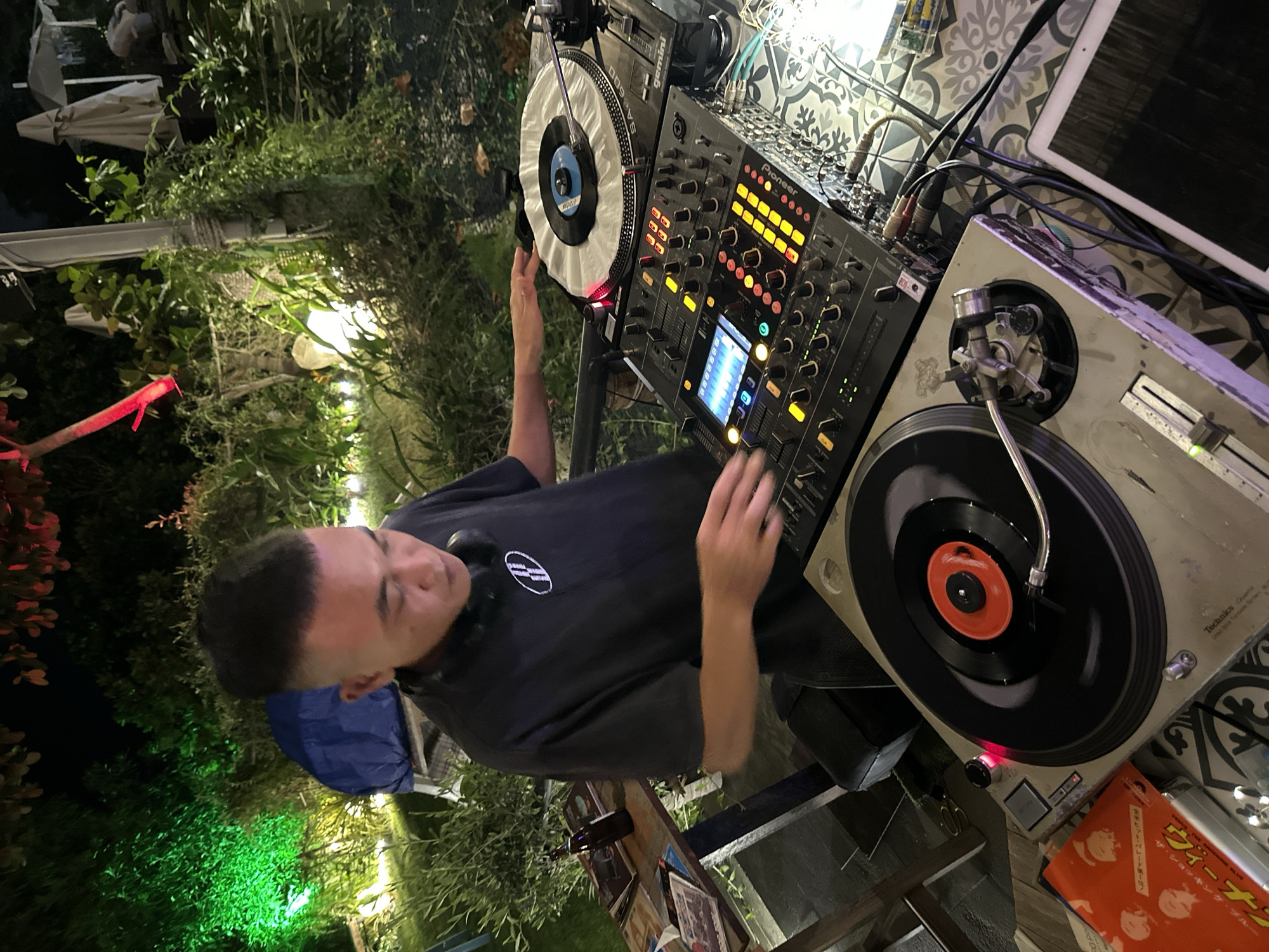 A person is shown DJing outdoors at night in Mũi Né, Vietnam. The DJ, wearing a black t-shirt and headphones, is operating a Pioneer DJ mixer and two turntables. Green and red lights illuminate the surrounding lush vegetation, and several white umbrellas are visible in the background. The scene appears to be set in a garden or outdoor entertainment area.