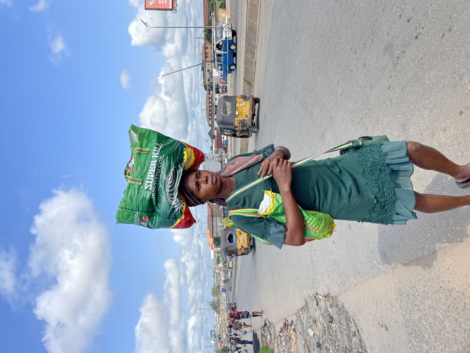 A Nigerian woman walks down a paved road in Fegge, Nigeria, carrying a large green bag labeled "SUPER KILL MOSQUITO COIL" on her head. She also has a smaller green bag slung over her shoulder. The woman is wearing a green dress and a patterned headscarf. The scene appears to be during the day, with a bright blue sky and scattered white clouds. Traffic, including cars and a yellow auto-rickshaw, is visible on the road behind her. The surrounding area shows buildings and some signs of urban life. The overall impression is of a busy street scene in Nigeria.