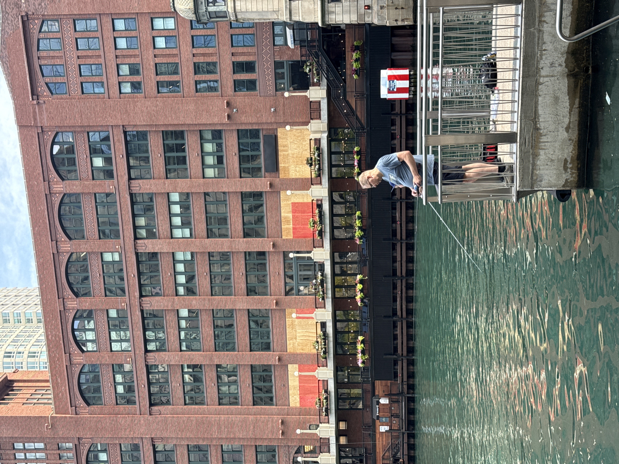 A man is fishing from a pier along the Chicago River in Chicago, Illinois. He is standing on a concrete pier with a metal railing, holding a fishing rod. The water is green and choppy, reflecting the red brick building behind him. The building has many windows and is several stories high. The man is wearing a blue t-shirt and black shorts. There is a red and white sign on the pier behind him. The scene appears to be a public area with the riverfront accessible for recreational activities.