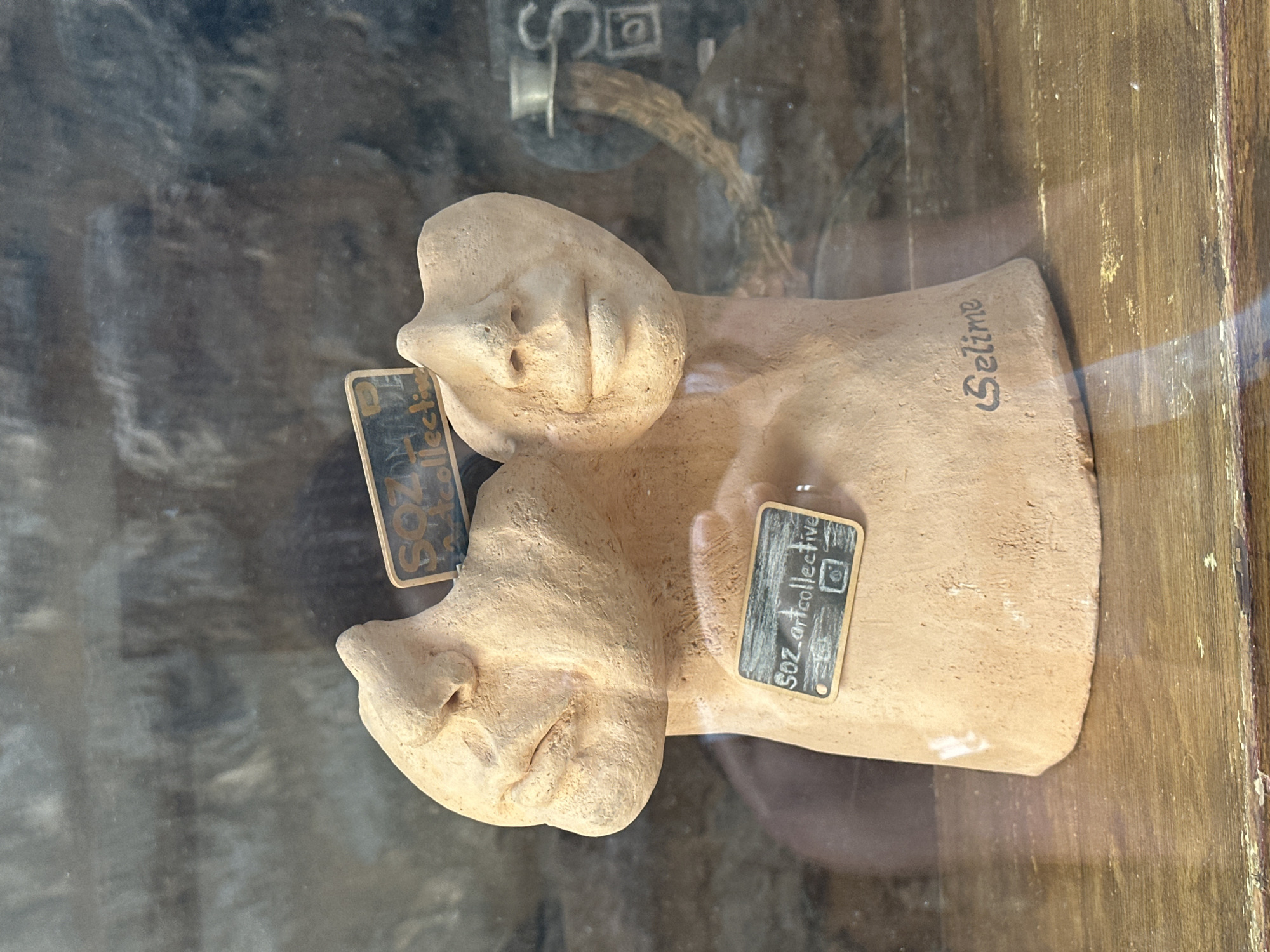 The image shows a terracotta sculpture of two conjoined heads with protruding noses, possibly displayed in a storefront in Budva, Montenegro. Small, hand-lettered black tags, presumably price or identifying labels, are affixed to the sculpture. One tag reads "SOZ art collective" and displays an Instagram logo. The name "Selime" is inscribed on the base of the sculpture. The sculpture is behind glass, with reflections visible. The background is blurry and hints at a textured, possibly wooden, surface.