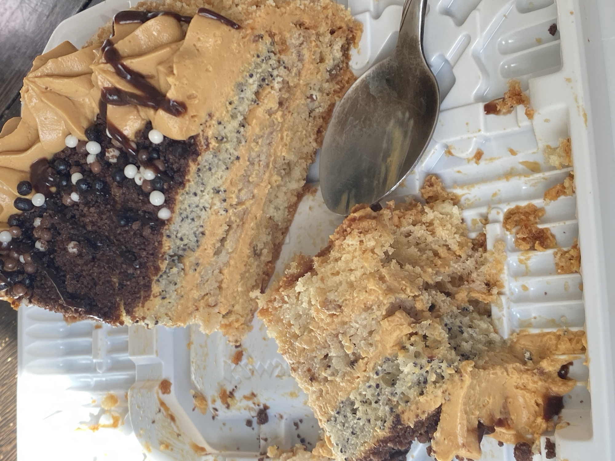 The image shows a close-up, top-down view of a slice of cake and a spoon in a white plastic container. The cake appears to be layered with a light-colored sponge cake and a caramel-colored frosting, with dark chocolate cake layers and a topping of chocolate sprinkles and small white balls. A portion of the cake has been eaten, and crumbs are scattered within the container. The spoon is metallic and positioned to the right of the cake slice. The background is out of focus and appears to be a wooden surface, suggesting the container is on a table. No specific location in Novokuznetsk, Russia can be identified from the image.
