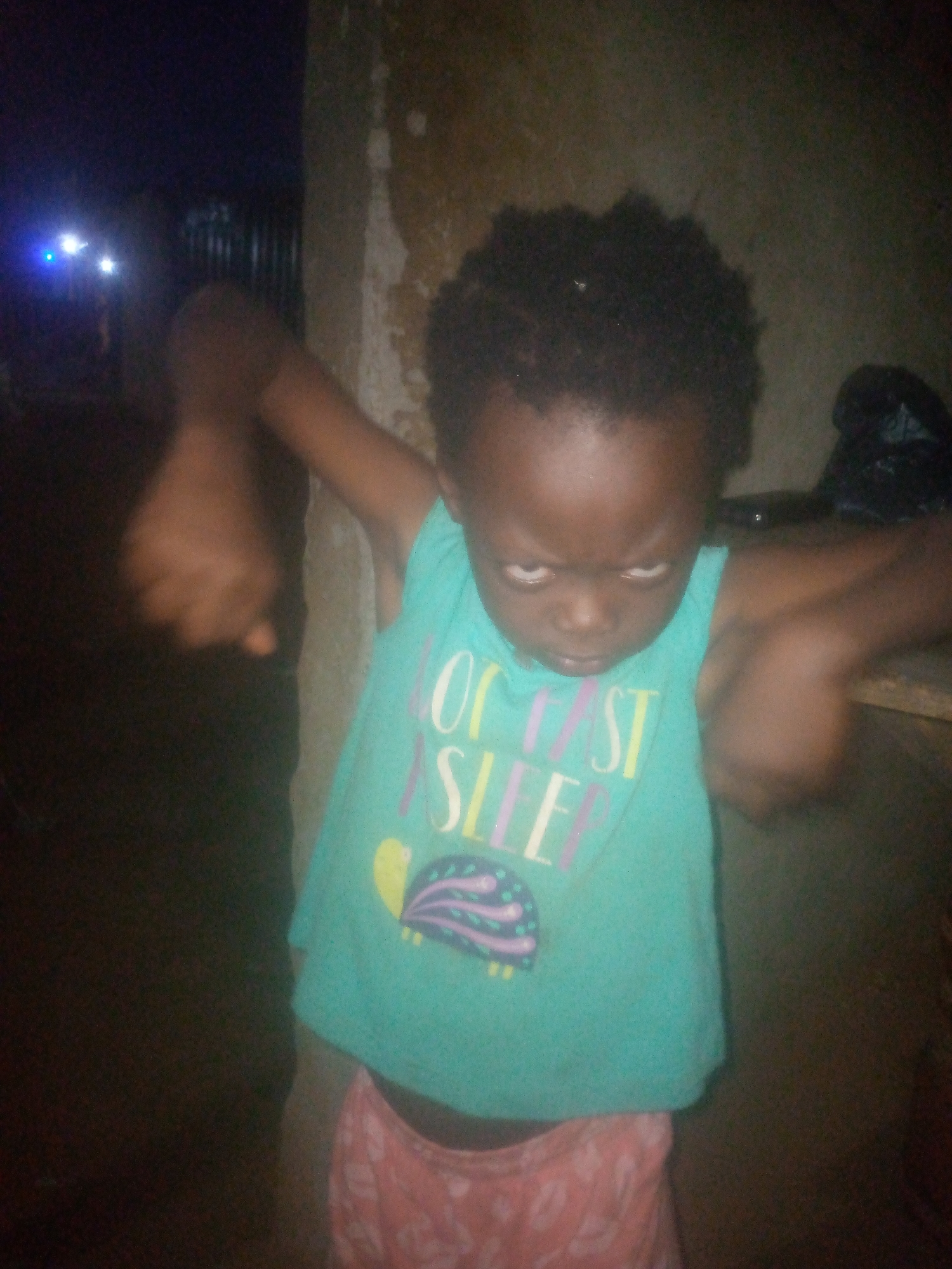 A child with dark skin and short, dark curly hair is standing in front of what appears to be a wall. The child is wearing a light blue sleeveless shirt with the words "OF COURSE I SLEEP" printed on it, along with a turtle image. The child is also wearing pink patterned shorts. The child's arms are outstretched, and their face appears to be contorted, as if angry or in distress. The location is the city of Unknown, Unknown. The background is dark, with a blurry area that includes some faint lights in the distance. The overall lighting is poor, obscuring details of the surroundings.