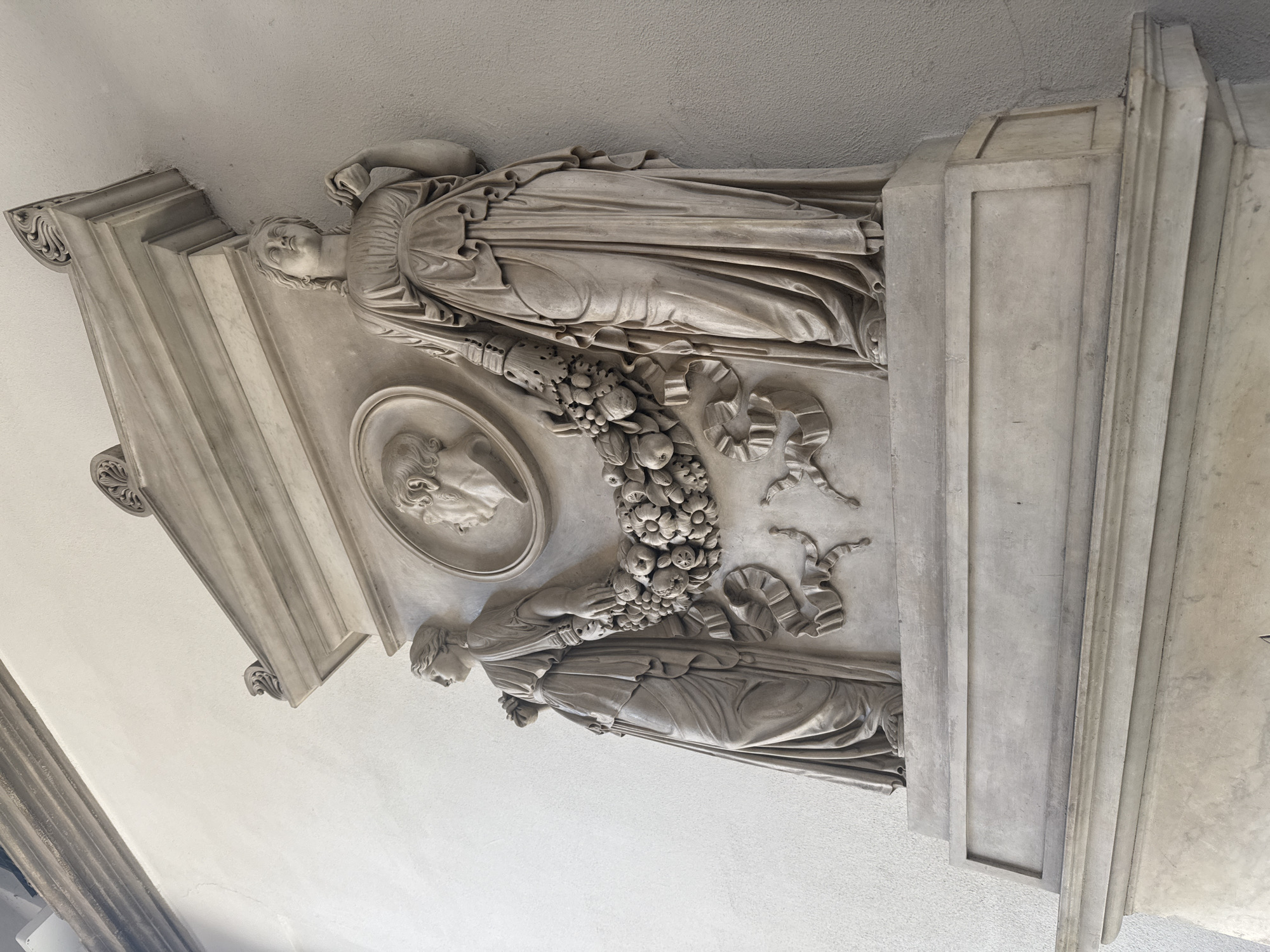 The image shows a marble relief located in Milan, Italy, specifically at the colonnade of the Ospedale Maggiore (now the State University). The relief is situated on a wall or pillar of the colonnade. It features two reclining figures of women in classical drapery. Between them, at the center of the relief, is a circular medallion containing a male profile. A garland of fruits and ribbons connects the two figures, running between them and the medallion. The relief is framed by architectural elements.