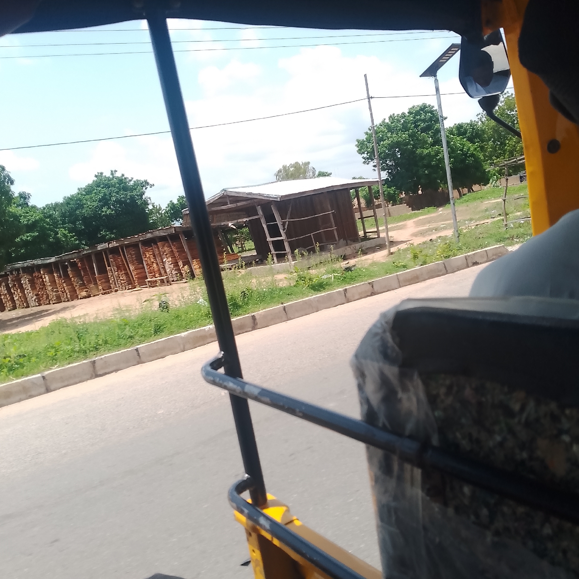 The image depicts a scene from Jalingo, Nigeria, viewed from inside a yellow vehicle, likely an auto-rickshaw. The foreground is dominated by the vehicle's interior structure, including black metal bars and a seat covered in dark fabric.
The scene outside reveals a roadside with a paved road and a concrete curb. To the left, a large structure holds stacks of lumber, organized in rows and leaning against supports, suggesting a lumberyard or construction site. Beyond the lumber, a rustic wooden shelter with a corrugated metal roof stands. The shelter appears partially open and has a ladder leaning against it.
In the background, lush green trees form a dense foliage, indicative of a tropical or sub-tropical environment. A solar-powered streetlamp is visible on a pole, and power lines stretch across the sky. The sky is a bright blue with scattered white clouds, suggesting a sunny day. The overall impression is a glimpse of daily life in a semi-urban or rural area of Nigeria, with evidence of commerce and infrastructure. The lighting and clear sky suggest it is daytime, likely late morning or early afternoon. There is no visible text in the image.