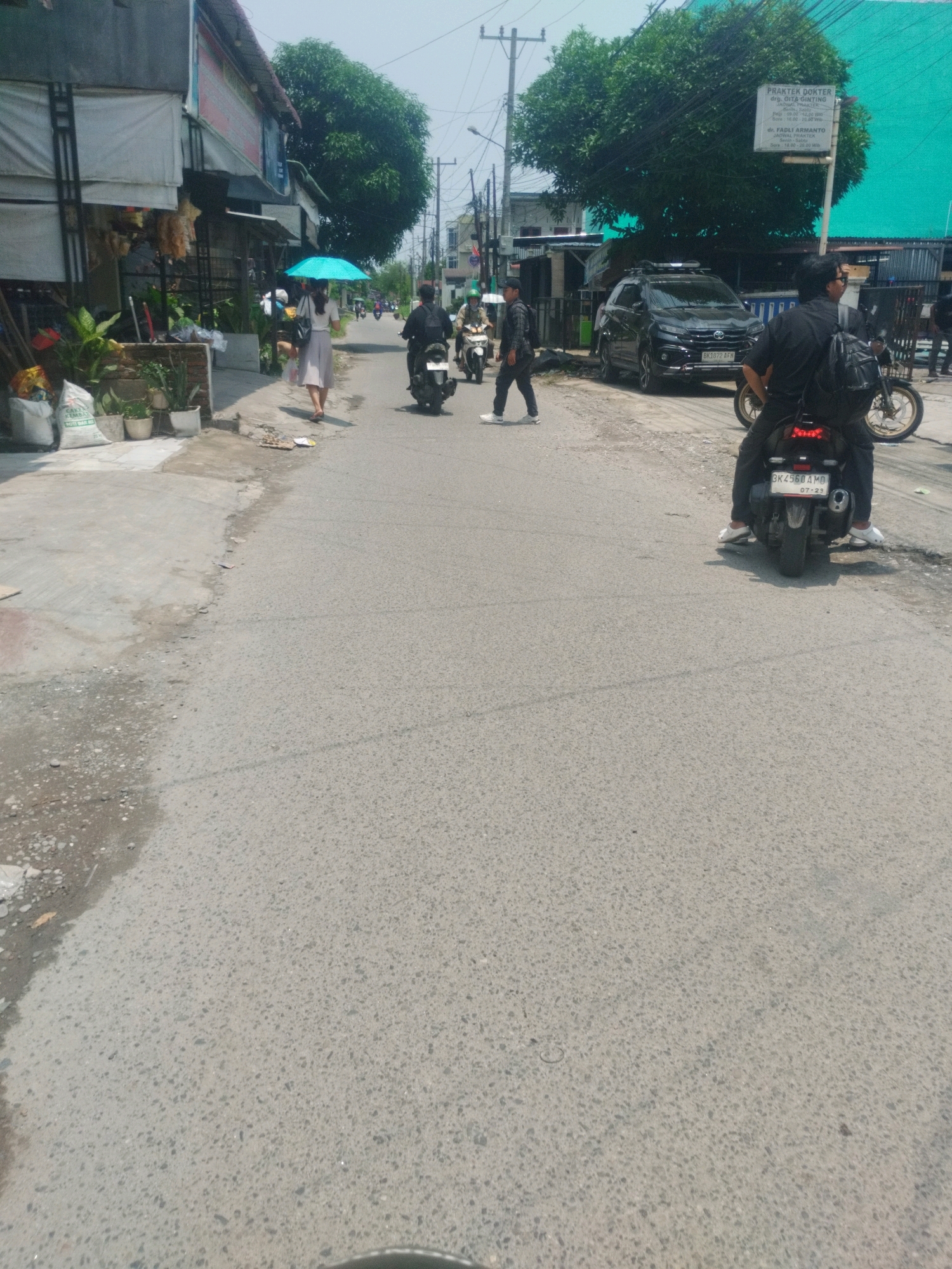 The image depicts a street scene in the City of Medan, Indonesia, on a sunny day. In the foreground, a person is riding a motorcycle from behind, wearing a black shirt and dark pants, with a black backpack. The motorcycle is dark-colored with a license plate reading "BK 4560 AMD". To the left, a woman with a blue umbrella walks away from the camera, wearing a white shirt and a light-colored skirt. She is walking on a sidewalk that has several potted plants and bags of material. In the middle ground, several other motorcycles and a black SUV are visible. The SUV has a license plate that reads "BK 1872 AFM". A sign with text in Indonesian is mounted on a pole near the SUV. The background shows buildings, trees, and utility poles, indicating an urban environment. The street is paved with asphalt, and there are some scattered debris on the side of the road. The overall atmosphere suggests a typical day in a bustling Indonesian city.