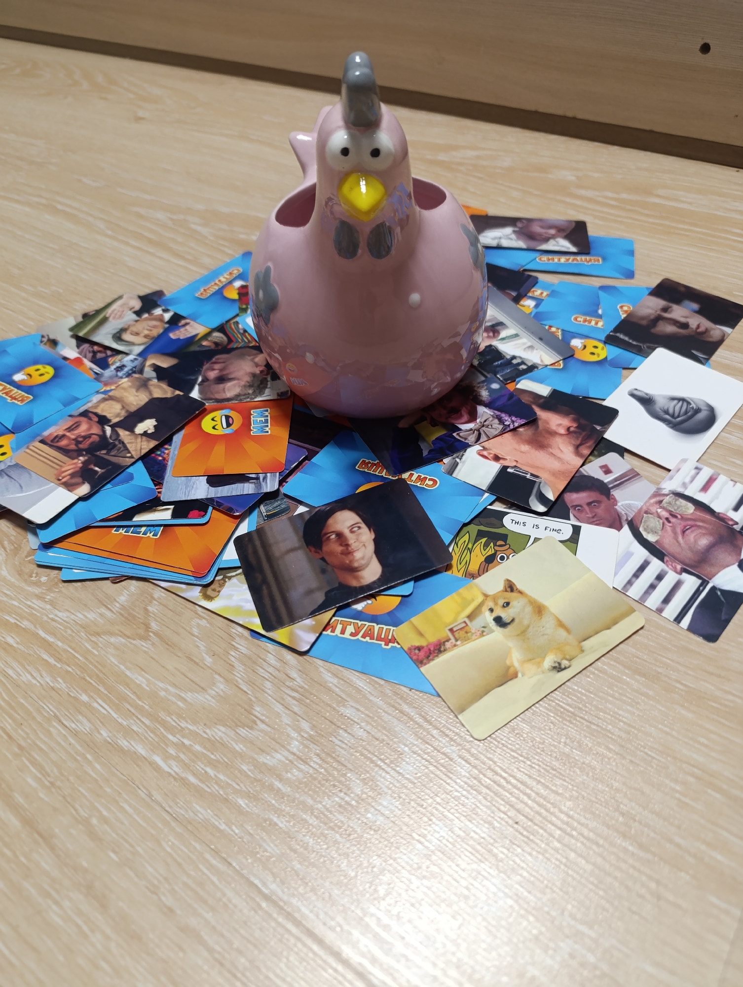 The image shows a pink ceramic chicken figurine placed on a pile of playing cards scattered on a wooden surface. The cards feature various images, including memes, celebrities, and what appear to be game-related graphics. Some of the text visible on the cards is in Russian, such as "МЕМ" (MEM) and "СИТУАЦИЯ" (SITUATION). The lighting suggests an indoor environment, possibly a room with wooden flooring and a wall in the background. There are no discernible geographic markers in the image that would indicate its location in Zlatopil, Ukraine. The scene is static and does not depict any actions or interactions.