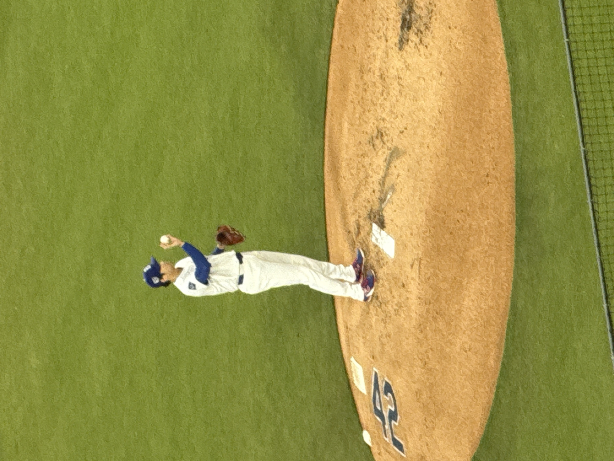 The image depicts a baseball pitcher, presumably of the Los Angeles Dodgers, mid-pitch on the pitcher's mound. He is wearing a Dodgers uniform with the number 42 prominently displayed on the mound. The environment includes the pitcher's mound, infield grass, and a portion of the stadium netting. The location is likely Dodger Stadium in Los Angeles, California, United States.