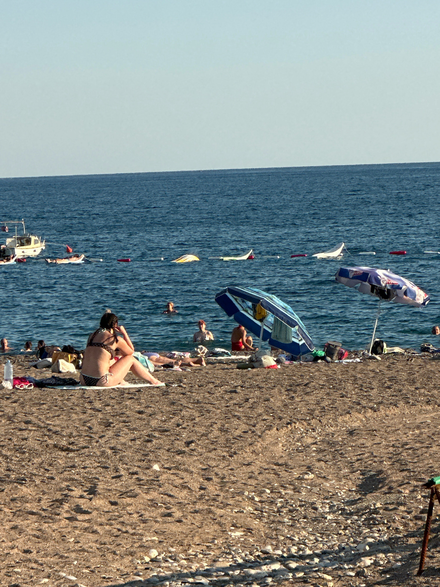 The image depicts a crowded beach scene with people sunbathing on the sand and swimming in the sea. In the foreground, a woman in a bikini is sitting on a towel, facing away from the camera. Several beach umbrellas are set up, providing shade for beachgoers. In the background, a small boat is anchored in the water, and people are visible in the sea. The beach appears to be covered in sand and pebbles, with the clear blue sea extending to the horizon under a bright sky. The scene suggests a typical summer day at a popular seaside resort.