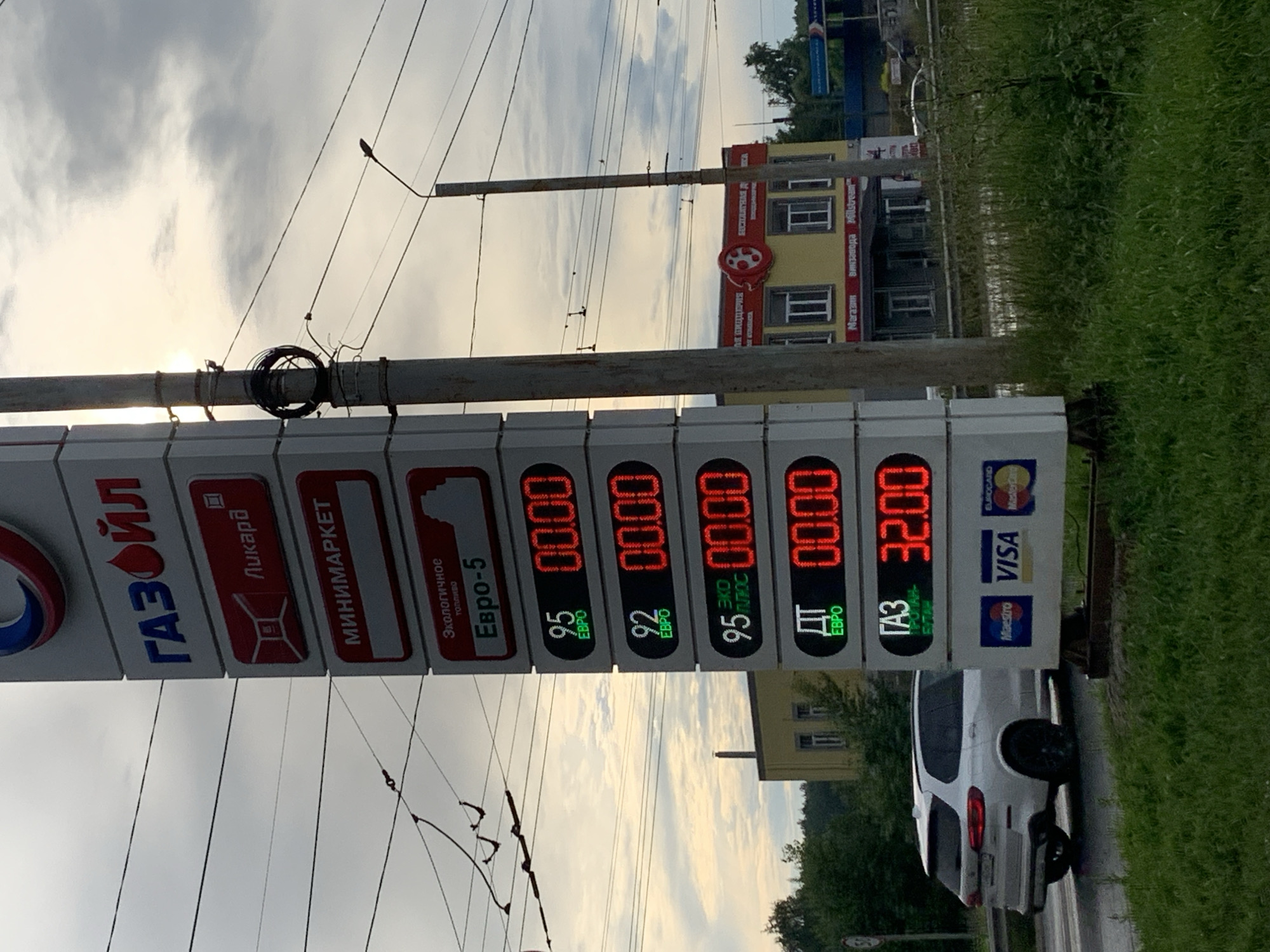 This media file captures a street-level view of a gas station with a prominent price display board. The sign features the "ГАЗОЙЛ" (Gazoyl) logo and lists various fuel types and their prices, including "Евро-5" (Euro-5) and "Газ" (Gas). The prices are shown in digital format, with some reading "00.00" and one displaying "32.00". Below the fuel prices, payment method logos such as Visa and Mastercard are visible.
In the background, a yellow building with windows and signage suggesting a "ПИЦЦЕРИЯ" (Pizzeria) and a "Магазин" (Shop) is partially visible. The scene is set outdoors, with a cloudy sky suggesting it might be a late afternoon or early evening. The presence of overhead wires indicates an urban or semi-urban environment, possibly near a road where a white SUV is parked. The overall impression is that of a typical roadside service area in Russia, as indicated by the location context of Kokhma, Russia. The lighting is somewhat diffused due to the overcast sky. There are no people clearly visible in the frame, focusing the attention on the gas station infrastructure and the surrounding buildings.