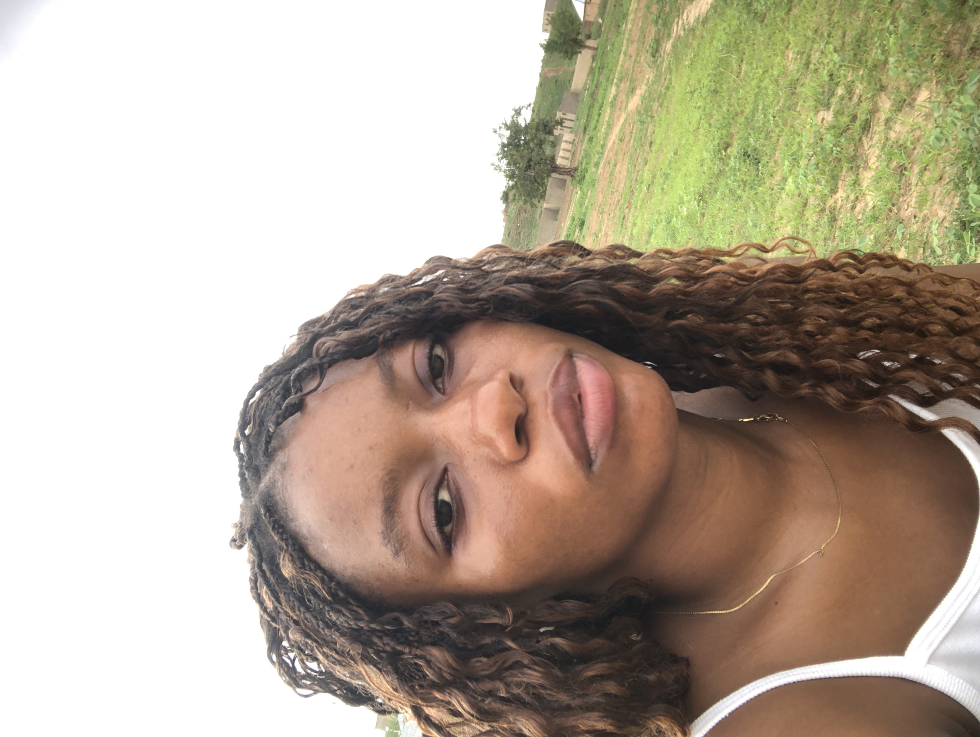 A medium-angle close-up shot of a dark-skinned woman with brown curly, braided hair wearing a white tank top and a delicate gold necklace. The background features a grassy field with some trees and what appears to be buildings in the distance. The sky is mostly white, indicating an overcast day. The picture was taken outdoors in Kashere, Nigeria.