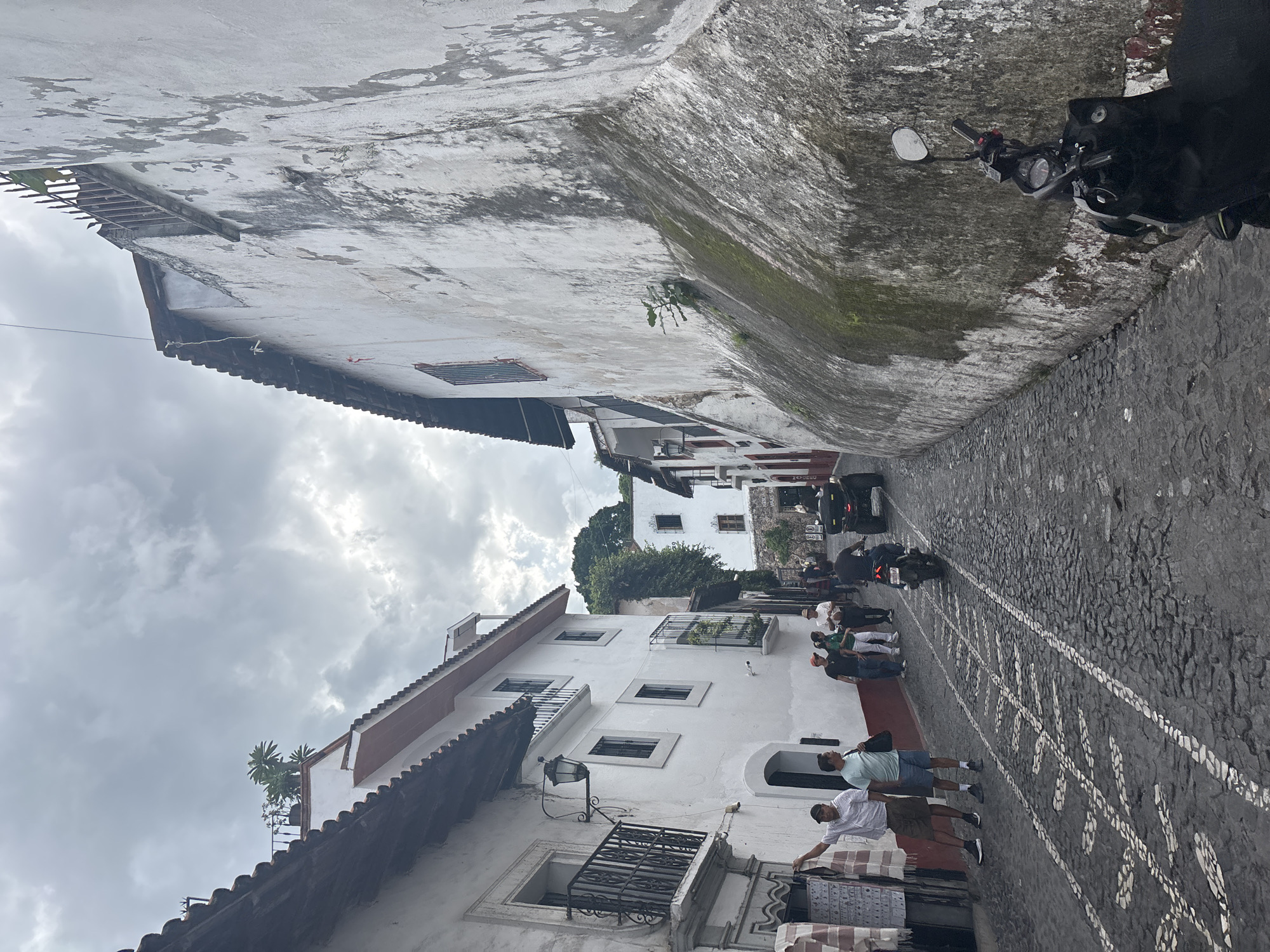 In Taxco, Mexico, a narrow cobblestone street winds uphill between whitewashed buildings with red-tiled roofs. The architecture features colonial-era balconies with wrought-iron railings, arched doorways, and decorative window grates. Several people are walking along the street, some browsing goods displayed outside a shop on the left. A motorcycle and an ATV are also present, suggesting a mode of transportation within the city. The sky is overcast with dramatic clouds, and lush green trees are visible in the background, hinting at the city's mountainous terrain. The overall scene captures the charm of a historic Mexican town.