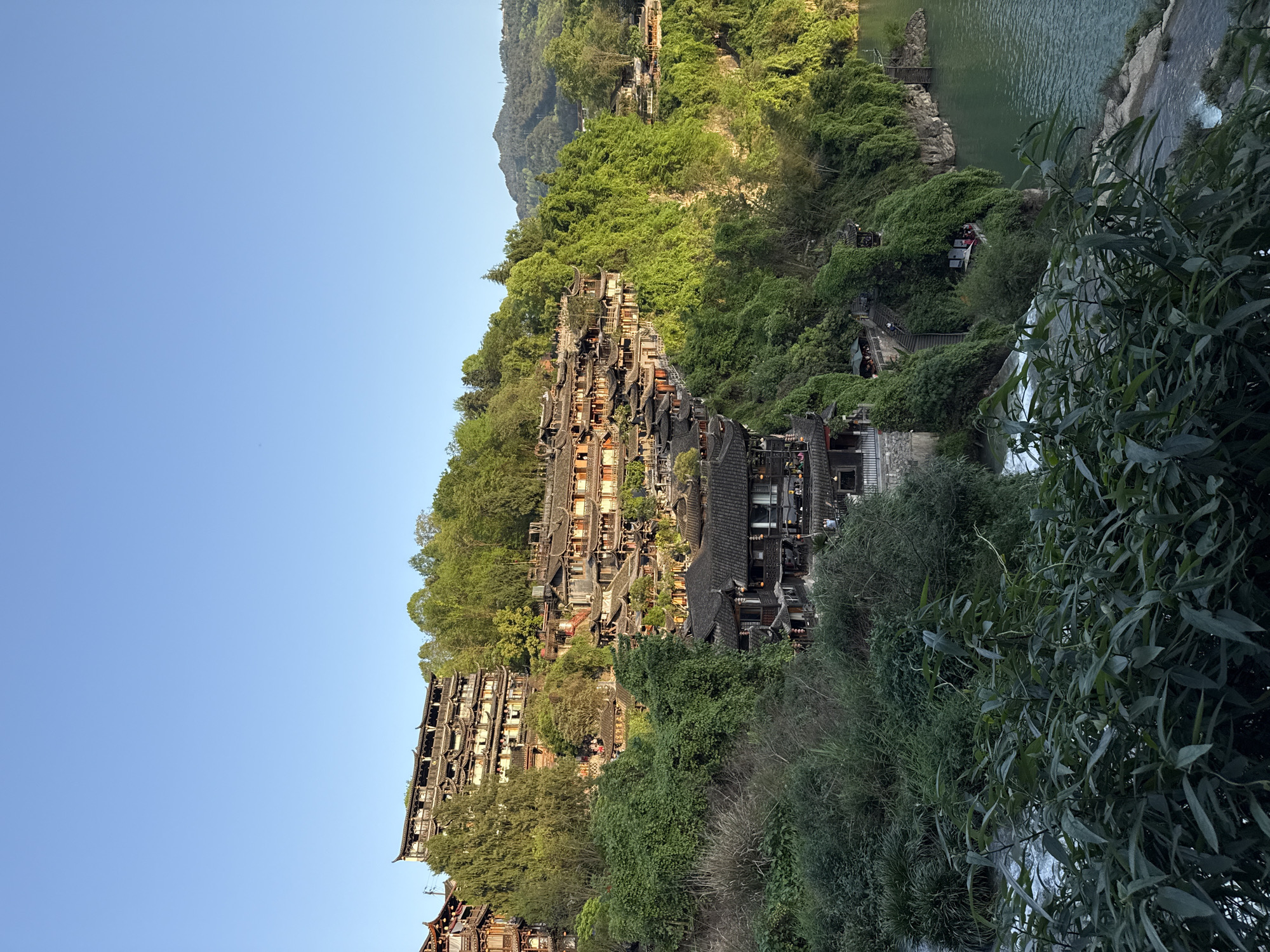 The image shows a hillside village in Furong, China. Traditional Chinese architecture is visible with multi-tiered wooden buildings nestled into the lush green landscape. A river runs alongside the village, and a waterfall is visible partially obscured by vegetation. There is a pathway with people on it next to the buildings. The sky is clear and blue.