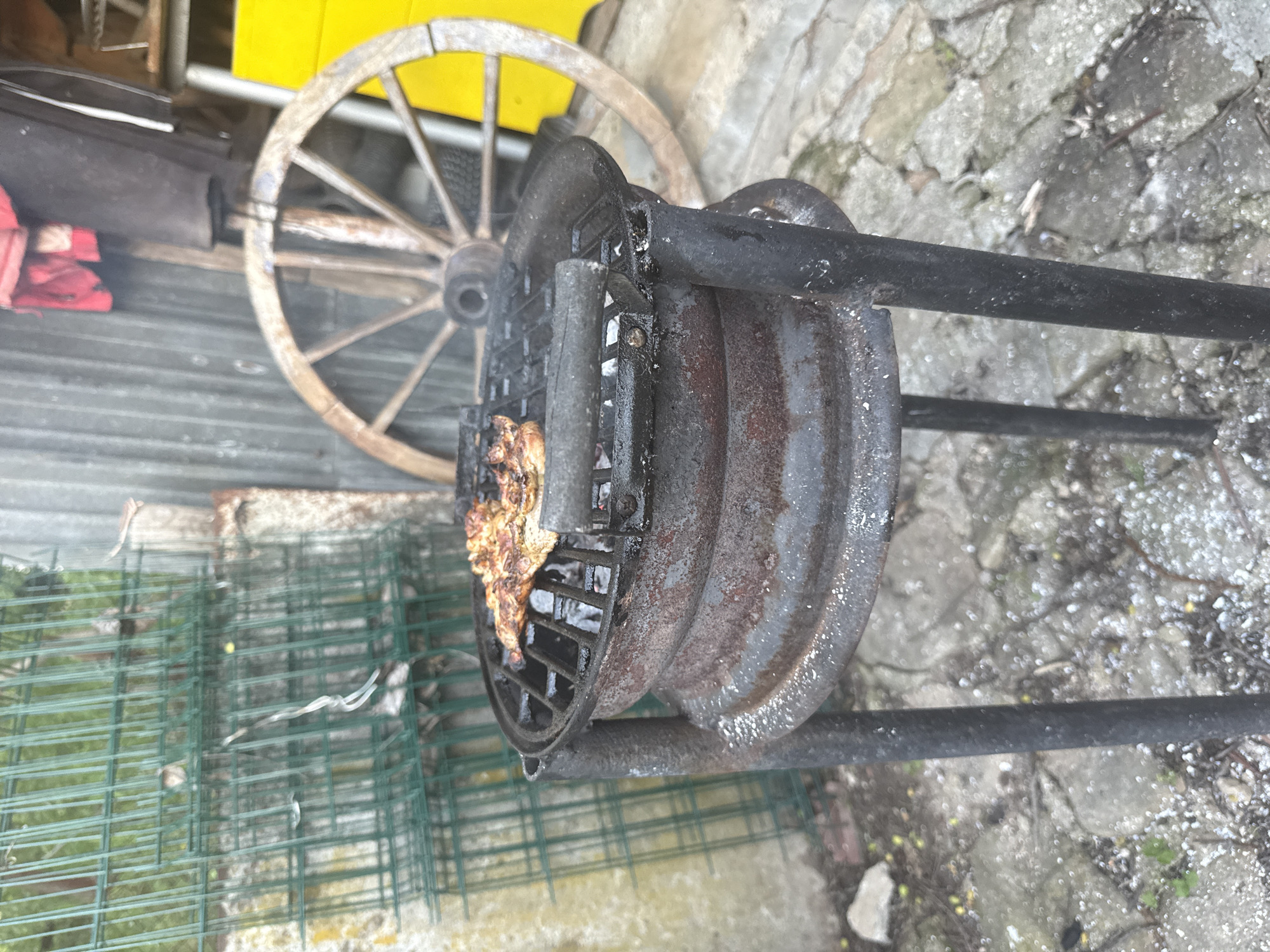 The image depicts a piece of grilled meat cooking on a rustic, black metal grill. The grill is positioned outdoors on a concrete or stone surface, next to a green wire fence and a wooden wagon wheel. The surrounding environment shows signs of wear and age, suggesting a backyard or outdoor cooking area in the countryside. The location in the city of Boltata, Bulgaria is noted, but no elements in the image directly confirm this. There are no people visible in the frame.