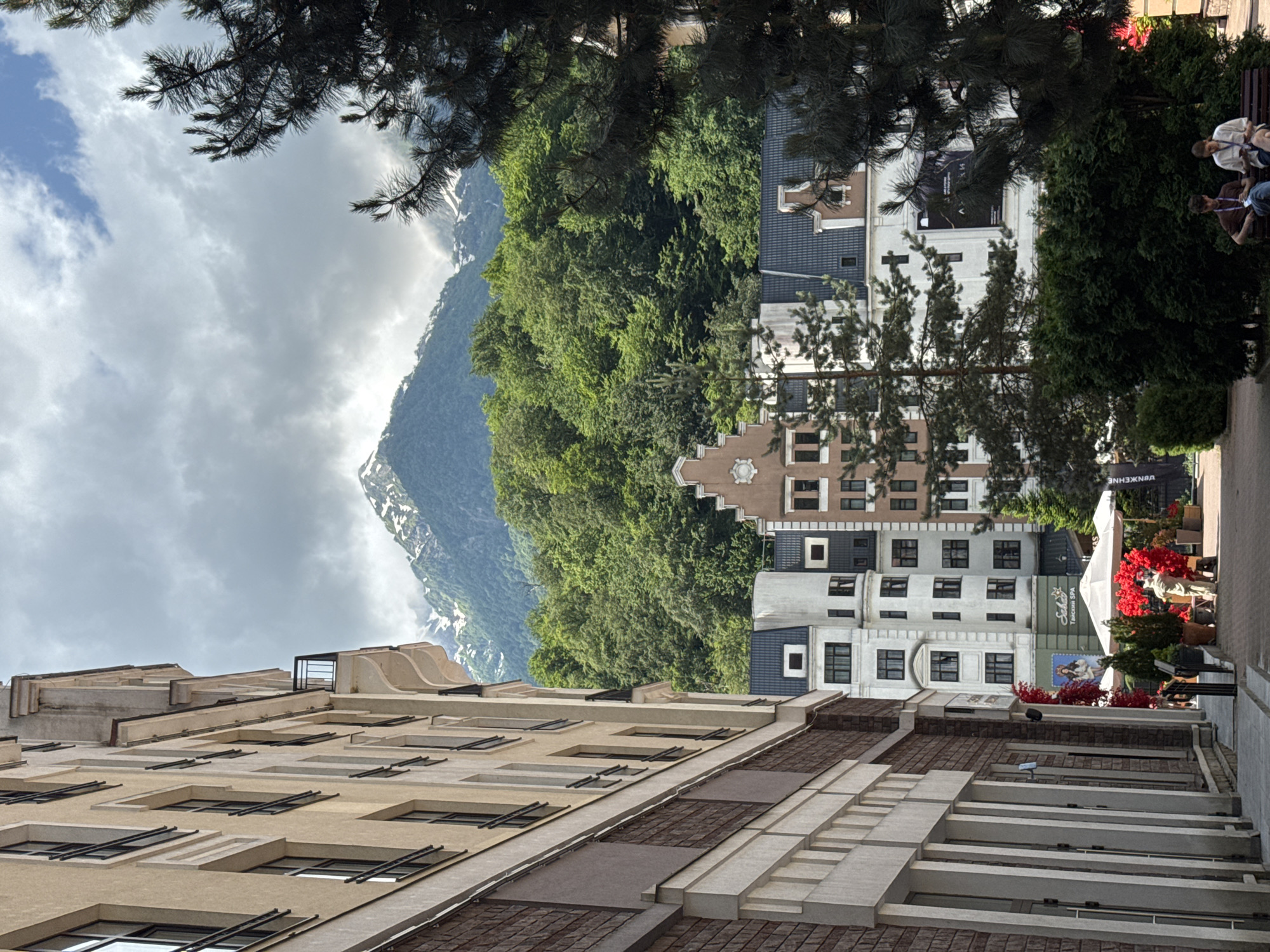 The image depicts a scene in Estosadok, Russia, characterized by a juxtaposition of European-style architecture and a mountainous, forested backdrop. In the foreground, a multi-story building with a beige and brick facade runs along the left side of the frame, featuring multiple windows and balconies. To the right, a streetscape unfolds with a row of buildings exhibiting diverse architectural styles, including a white building with a cream-colored gable and a brown building with decorative detailing. A variety of trees and lush green foliage are visible throughout the scene, with a prominent evergreen tree in the mid-ground and darker pine trees to the right. Beyond the buildings, a dense forest covers a mountain slope, which is capped with snow, suggesting a high-altitude or seasonal environment. The sky is partly cloudy, with patches of blue and significant cloud cover. In the lower right portion of the image, two individuals are seated on a bench, appearing to be engaged in conversation or reading. A person in a light-colored outfit is standing near a display of red flowers and potted plants in the mid-ground, under a white umbrella.
