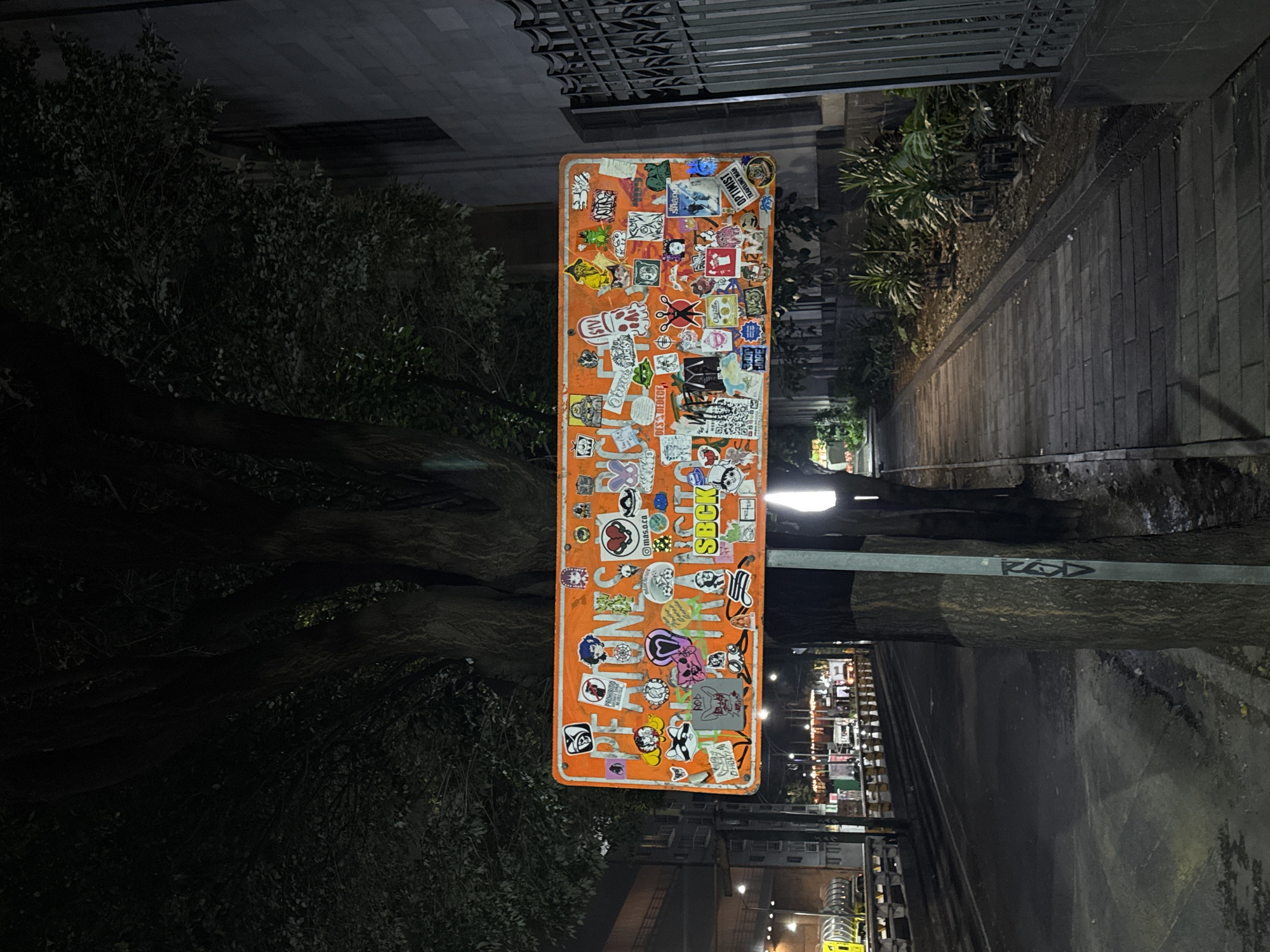 The image, rotated clockwise, captures a nighttime urban scene centered around a tall, narrow orange sign heavily adorned with numerous stickers. The sign is attached to a metal pole, which also features graffiti reading "ROD". The stickers on the sign display a variety of graphics and text, including phrases like "OPTIMIST TRAVELING MAN", "PROHIBIDO", "SBCK", "mas.o.ca", "DES BEDECE", "JOPE", "Lucky", "Chiülle", and "Shana".
To the left of the sign, a dense cluster of dark green foliage from a bush or small tree is visible, with a tree trunk peeking out from behind. On the right, a paved sidewalk or pathway made of dark grey blocks stretches into the distance, bordered by sparse low-lying greenery and another tree trunk. In the background, artificial lights illuminate what appears to be a street with buildings, suggesting a bustling environment despite the dark sky. Above the sign, part of a building facade with a decorative metal grate is visible. The scene is quiet, with no explicit activity, and the overall impression is that of an urban street at night.