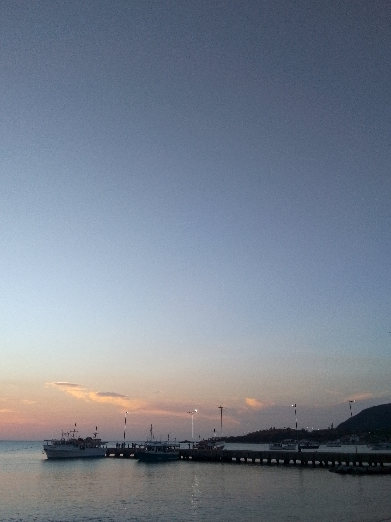 The image presents a serene twilight scene at a harbor, consistent with the coastal town of Juan Griego, Venezuela. A long pier stretches into the calm water, serving as a docking point for several boats. On the left, a larger white multi-deck boat is moored, while a smaller, darker vessel is docked further along the pier to the right. The pier is illuminated by several tall lampposts, casting bright white-yellow lights that reflect on the water's surface.
Numerous people are visible on the pier, mostly as dark silhouettes, seemingly strolling or standing, enjoying the evening ambiance. The sky dominates the upper portion of the frame, showcasing a beautiful gradient of colors typical of a sunset. It transitions from a dark, deep blue at the top to lighter blues, purples, and warm oranges and pinks near the horizon, where a few faint, wispy clouds are present. Dark, silhouetted hills or mountains form the backdrop on the right side of the image, contributing to the tranquil setting. The low light conditions of dusk give the scene a peaceful and reflective mood. The image exhibits some digital grain, particularly noticeable in the expansive sky. There is no visible text in the image.