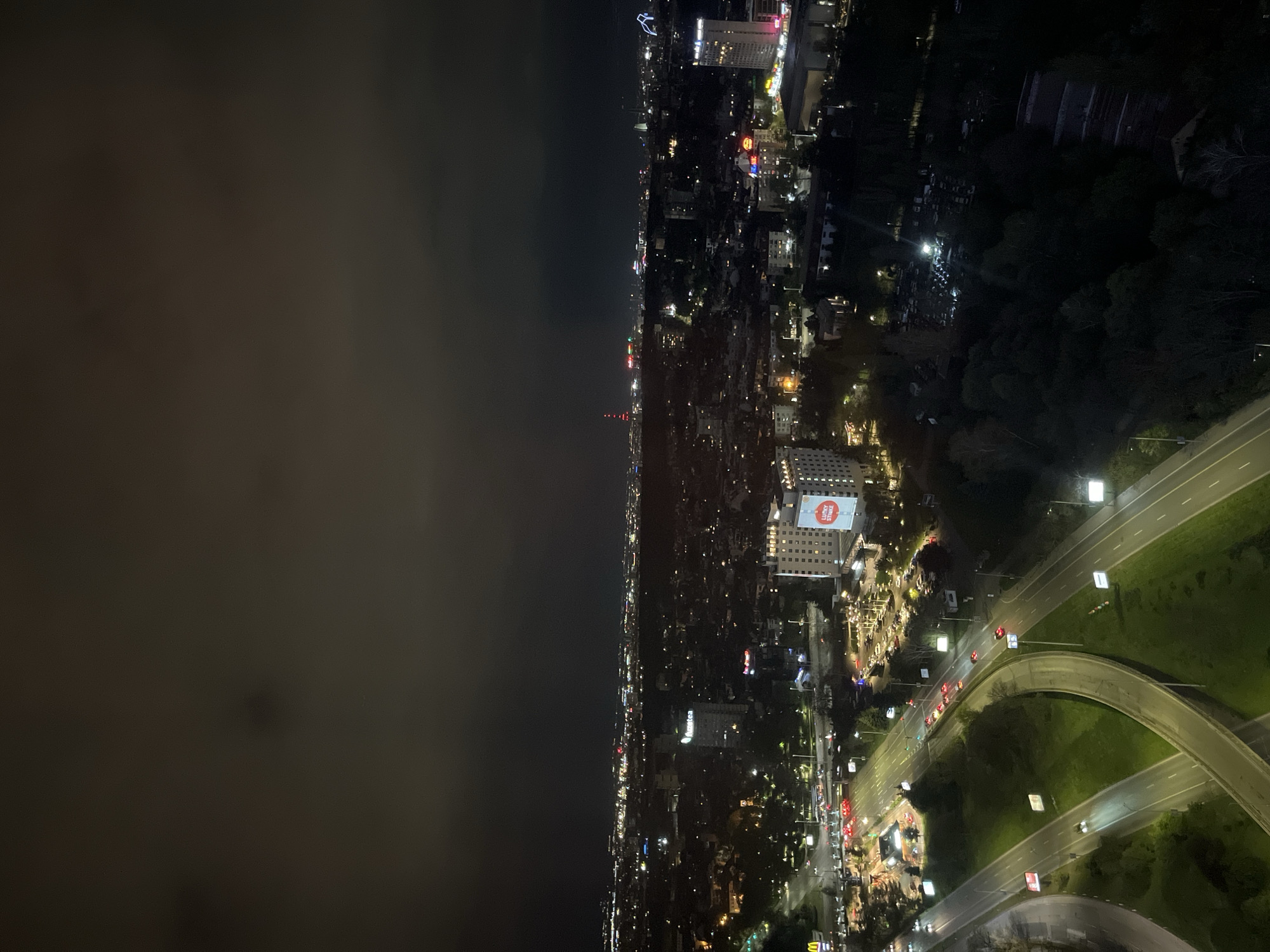 Here is a description of the image:
The image is a nighttime aerial view of a city, identified as Sofia, Bulgaria, based on distinctive landmarks. The right side of the frame shows illuminated highways and overpasses with visible traffic. Adjacent to the roadways is dense urban greenery, interspersed with lit buildings and streetlights. The central portion of the frame showcases a cityscape illuminated by numerous lights from buildings and street infrastructure, gradually receding into the distance. On one building is a prominent advertisement for "Lucky Strike". In the far distance is a radio tower. The left side of the image is dominated by an overcast or foggy sky, obscuring any possible features beyond the city's edge. The composition suggests that the photograph was taken from a high vantage point, possibly a tall building or a high elevation.