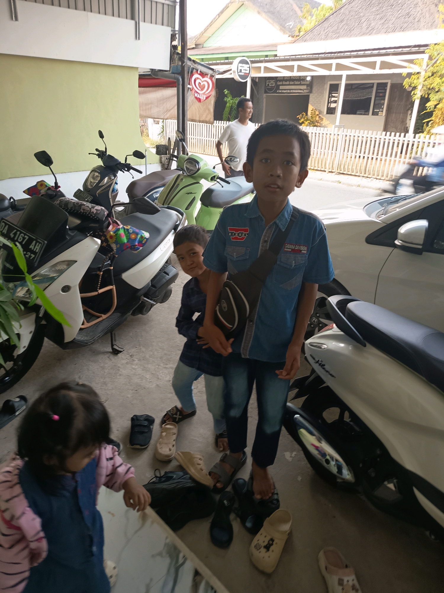 The image depicts an outdoor scene in Banjarmasin, Indonesia, likely in front of a building with a tiled roof and a pale green wall. Several motorcycles are parked, including a white Yamaha and a green scooter. In the foreground, a young girl in a pink and striped top and a blue jumpsuit is standing. Behind her, a boy in a blue denim shirt and jeans is looking towards the camera. To the left, another boy in a dark blue plaid shirt and jeans is partially visible. A man in a white t-shirt is standing in the background. Various footwear, including sandals and shoes, are scattered on the ground. A prominent "WALL'S" ice cream logo is visible on a red sign. The ground appears to be concrete.