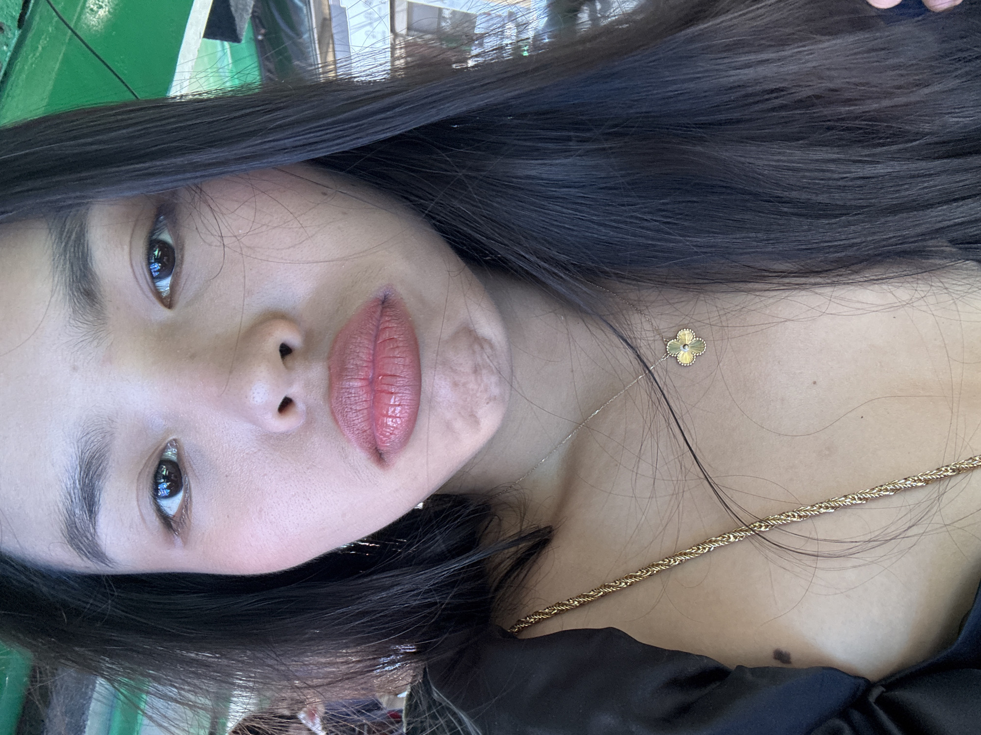 This is a close-up, portrait-style image of a woman, oriented on its side. The primary subject is the woman, who has long, dark hair that frames her face and extends over her shoulder. Her face is in a three-quarter view, looking directly towards the camera. She has prominent dark eyebrows and dark eyes, with visible reflections indicating bright ambient light. Her lips are painted with a reddish-pink matte lipstick, showing some texture. A small, raised mark or scar is visible on her lower cheek or jawline area.
She is adorned with two gold necklaces: a delicate chain featuring a small, four-leaf clover pendant with a central jewel, and a thicker, twisted rope-style chain. She is wearing a black garment, possibly a top or dress, visible around her shoulder and décolletage. Her skin appears fair.
The background is softly blurred, showing indistinct shapes and colors, including a noticeable green hue in the upper left, possibly from foliage or a structure. The lighting is bright and natural, suggesting a daytime setting. No specific activity beyond posing for the photograph is evident. There is no visible text in the image. The location context for this portrait is Davao City, Philippines.