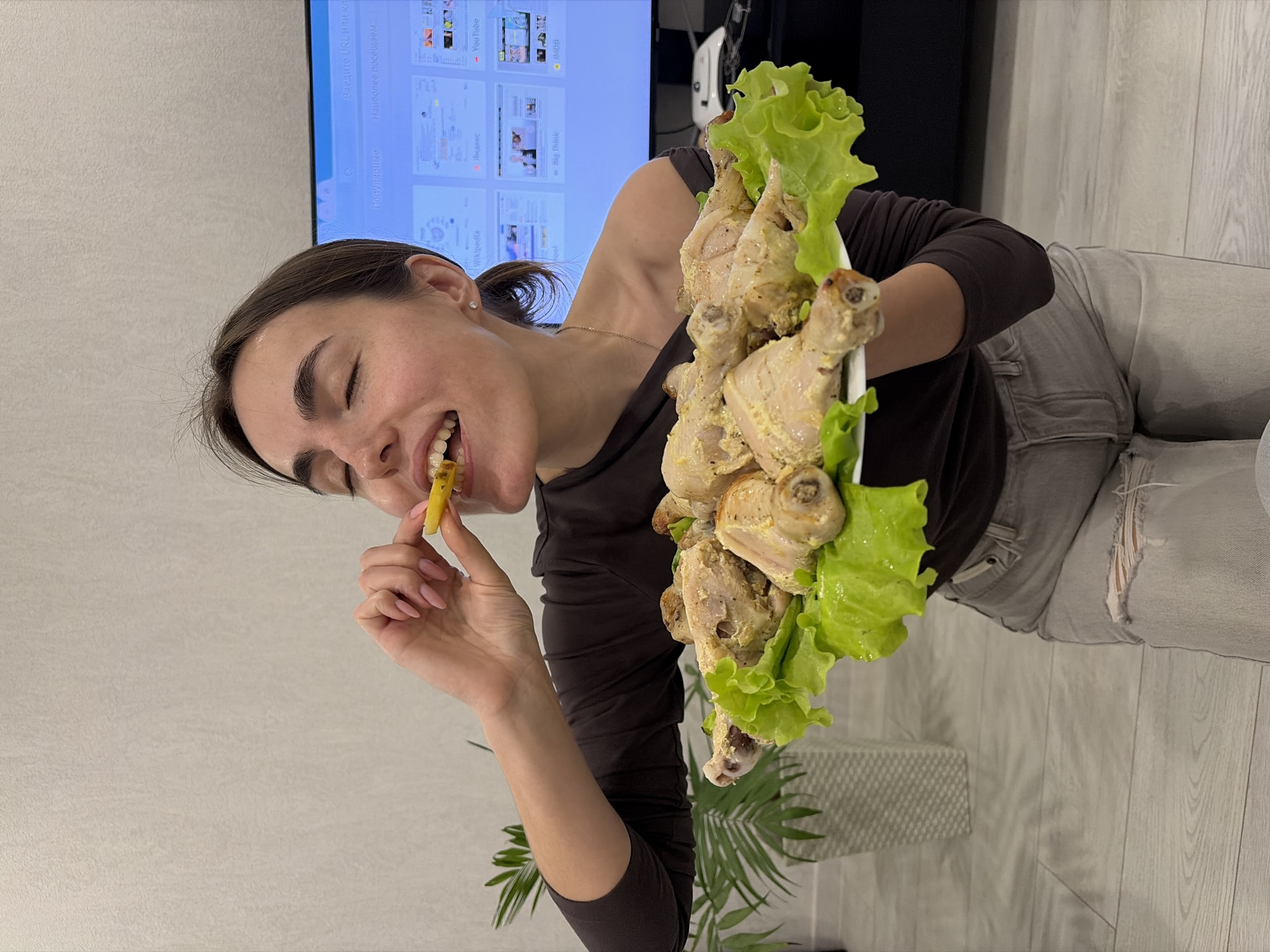 A woman with dark hair pulled back into a ponytail is holding a plate of cooked chicken legs garnished with lettuce. She is wearing a dark brown long-sleeved shirt and light grey jeans with ripped knees. She is holding a small yellow piece of food in her fingers and putting it in her mouth with her eyes closed. In the background, there is a television screen with website pages visible, and a black cabinet. The photo appears to be taken indoors, possibly in a home or apartment, with light grey floors and a faux palm plant in the lower portion of the image. There is no explicit information available in the image to confirm the location in Sarapul, Russia.