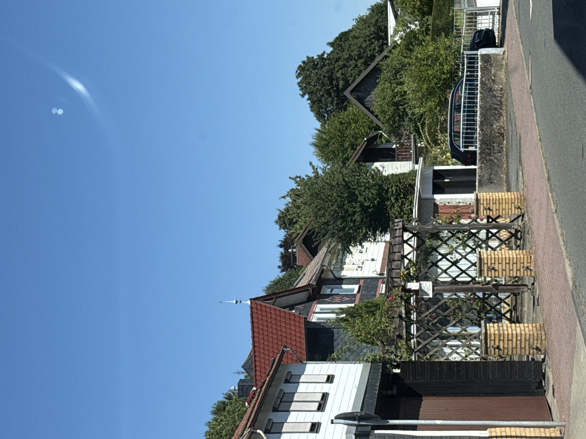 The image shows a residential street in Bad Sachsa, Germany, on a clear, sunny day. In the foreground, a sidewalk and road are visible. A row of houses with red tile roofs and white exteriors line the street. Trees and greenery are abundant, suggesting a suburban or semi-rural setting. A dark-colored car is parked behind a white metal fence in front of one of the houses. The sky is a vibrant blue with a faint white streak, possibly a contrail. The overall impression is one of a peaceful, well-maintained neighborhood.