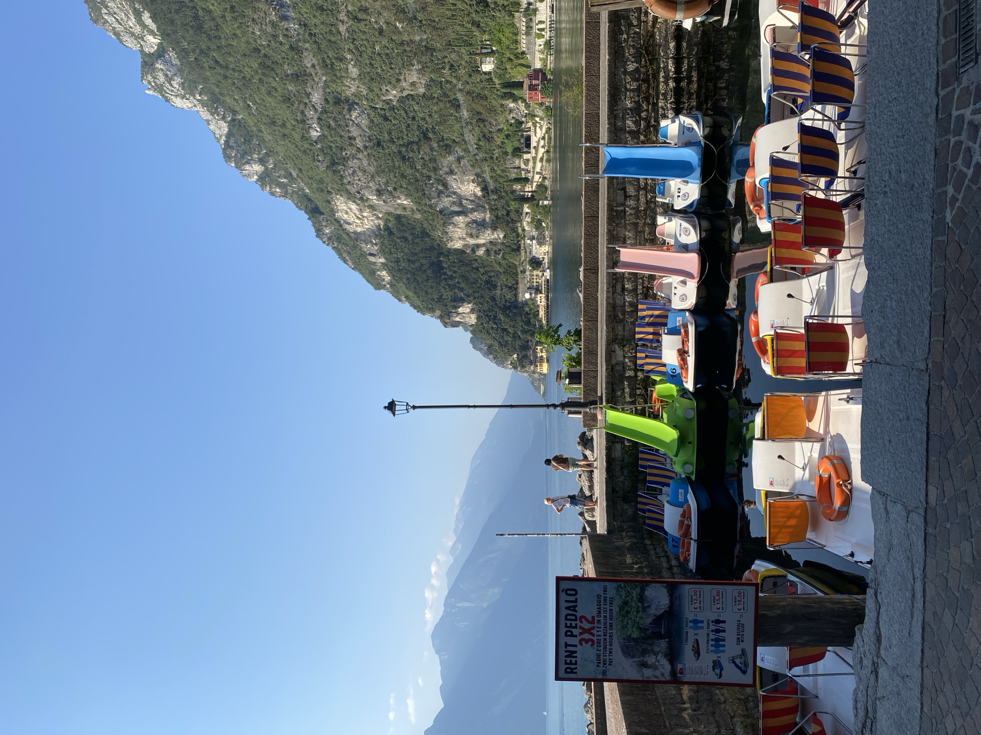 The image shows a picturesque scene in Riva del Garda, Italy, likely on a sunny day, with a clear blue sky and calm lake water. In the foreground, several colorful pedal boats are docked. Some are equipped with slides for children, and others have rows of striped chairs. A prominent sign on the left side advertises "RENT PEDALO 3X2", indicating a special offer for renting pedal boats.
Beyond the docked boats, the tranquil waters of Lake Garda stretch towards distant, majestic mountains. The shoreline is lined with a stone wall, and a lamppost stands tall near two individuals who appear to be tourists, looking out at the lake. The background features a charming town nestled at the base of a steep, green mountainside, with buildings visible along the waterfront and scattered amidst the trees. The overall atmosphere is one of leisure and natural beauty.