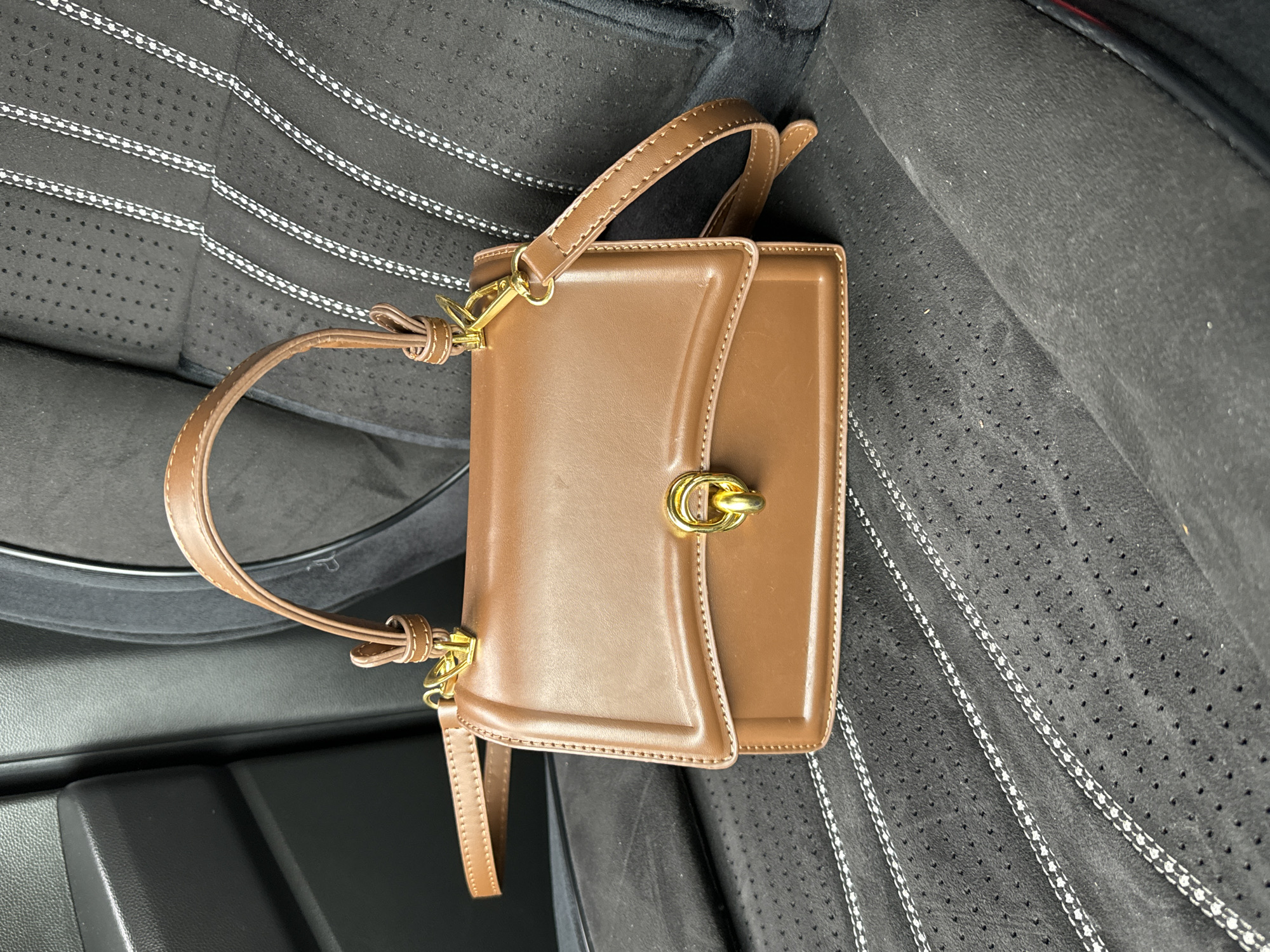 A medium shot captures a structured brown handbag resting on a dark car seat. The handbag, positioned diagonally across the frame, is made of a smooth, tan-brown material, likely leather or faux leather. It features a top handle attached by gold-colored metal rings and a front flap secured with prominent gold-toned hardware, possibly a twist lock. Visible stitching outlines the edges of the bag and its handle.
The car seat, serving as the background, is dark grey or black with a finely textured surface. It is adorned with multiple parallel lines of white dotted stitching, creating a distinctive striped pattern. Parts of the seat also appear perforated. To the left and bottom of the handbag, dark, non-reflective plastic elements of the car's interior are visible, likely parts of the door panel or center console. The lighting is soft and even, suggesting daylight filtering into the car. No people are present, and there is no discernible text. The image is set within a car in Chaikovsky, Russia.