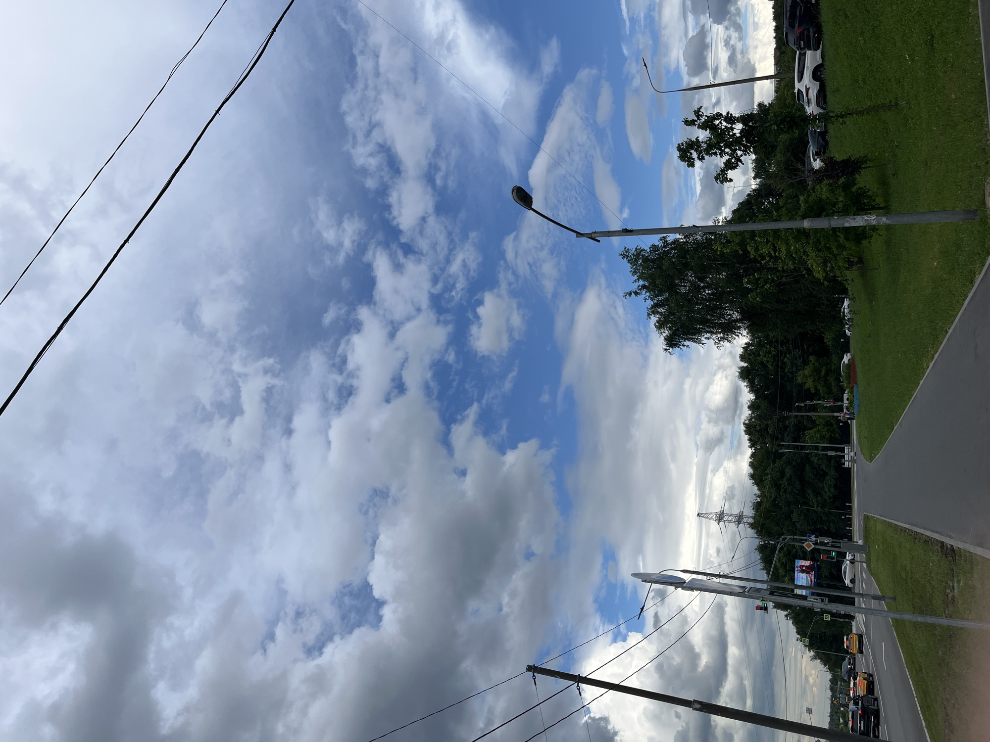 This is a photograph taken on a cloudy day, likely in the afternoon, of a street scene with a focus on the sky and overhead wires. The location is identified as Moscow, Russia.
In the foreground, a paved pedestrian walkway is visible, bordered by a grassy median. Beyond the median, a multi-lane road carries several vehicles, including cars and taxis. Traffic signals are present, indicating a regulated intersection. A tall metal pylon, likely a transmission tower, stands in the background, partially obscured by dense green trees that form a treeline along the edge of the road.
The sky dominates the upper half of the frame, filled with a dramatic mix of white and grey clouds against patches of bright blue. Several power lines stretch diagonally across the sky, supported by utility poles visible on the left side of the image. Streetlights are positioned along the side of the road. In the middle ground, there is a billboard visible featuring two individuals.
The overall impression is an urban landscape under a dynamic, partly cloudy sky. There are no people clearly depicted or actively engaged in any discernible actions within the frame. The scene is static, focusing on infrastructure and atmospheric conditions.