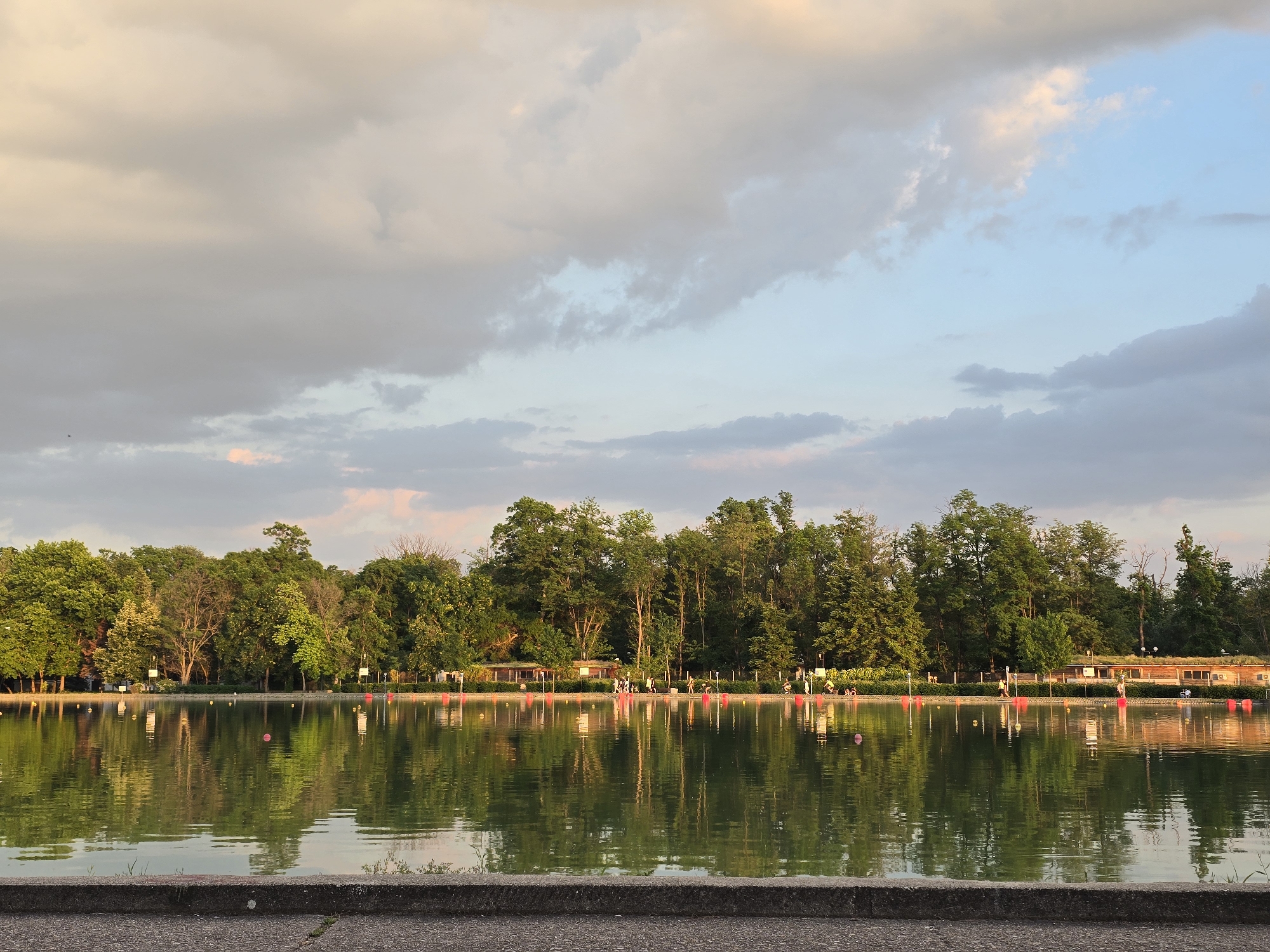 The image shows a lake in Unknown, Unknown. The water is calm and reflects the trees and sky. A row of trees lines the opposite bank of the lake. There is a building on the far right of the image with people walking in front of it. Red buoys float in the water. The sky is filled with clouds, and the lighting suggests it is late afternoon or early evening. A concrete barrier is visible in the foreground.